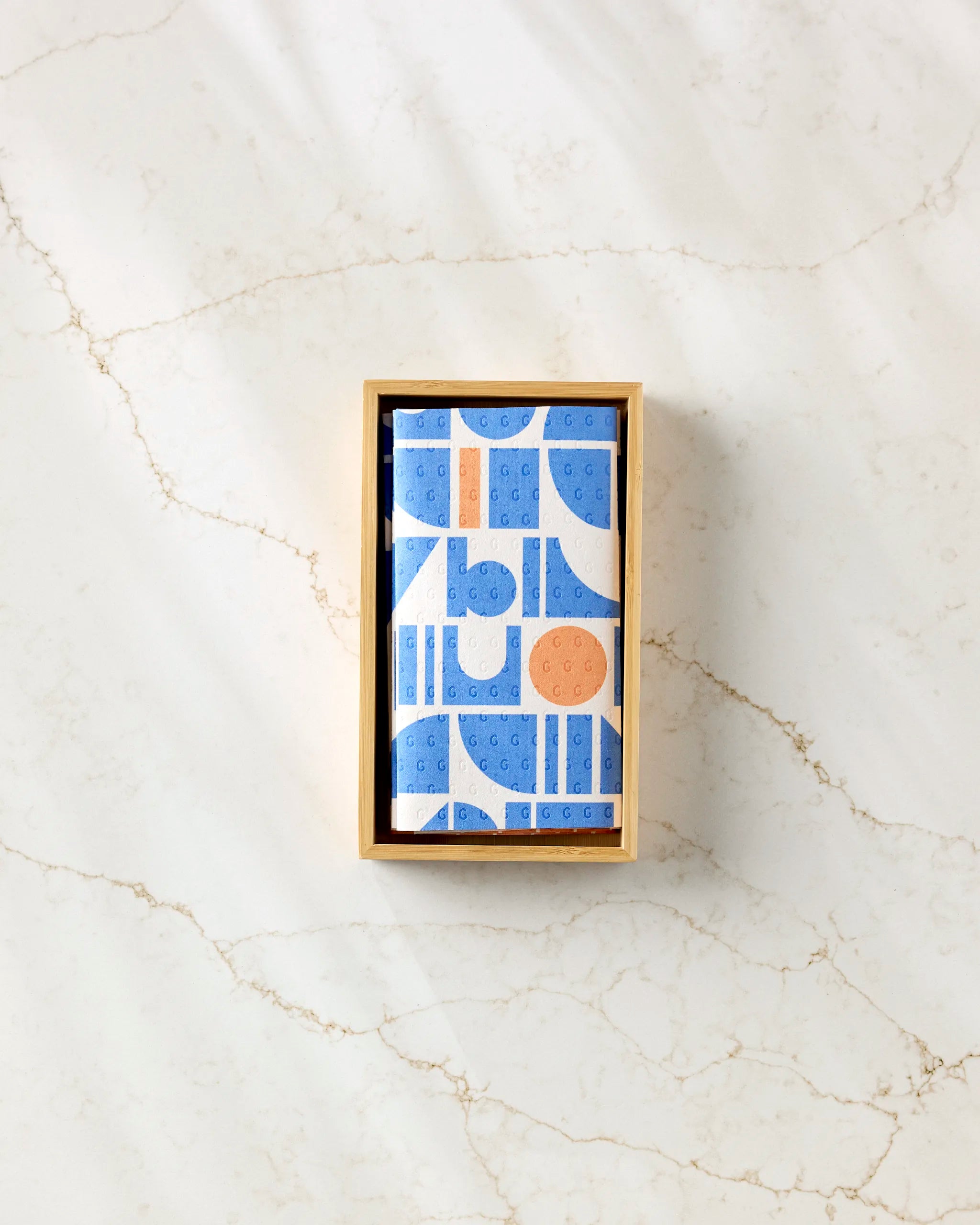 A rectangular Sun Beam Harmony box of eco-friendly kitchen towels by Happy Hemera, with a blue and orange geometric pattern, rests on a white marble surface with thin brown veining, viewed from above.