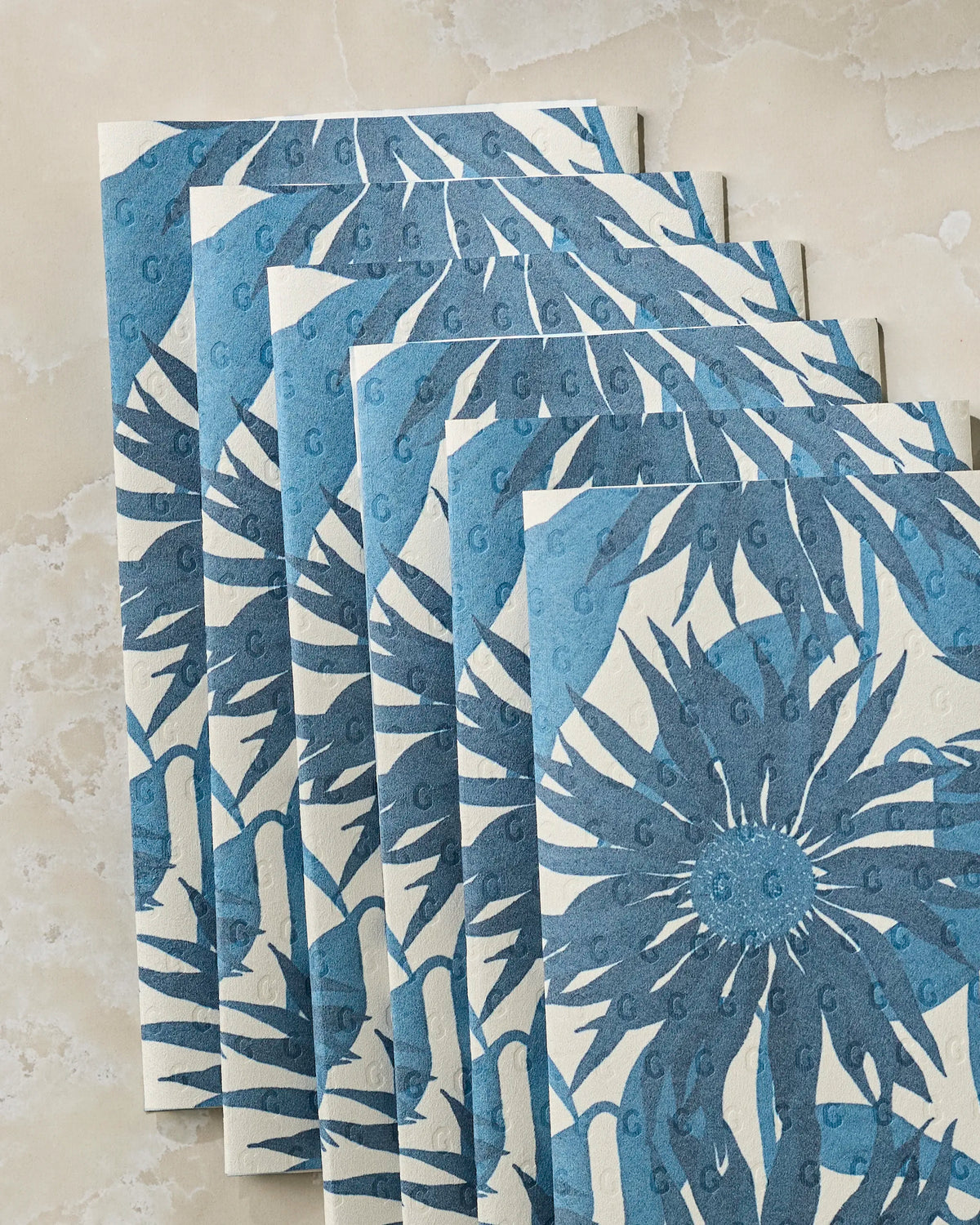 Six Sundara Frost cards by Geometry, displaying a blue floral pattern with overlapping edges, are fanned out on a marble surface. Each card features large petals radiating from circular centers on a light background, evoking the elegant look of eco-friendly kitchen towels.