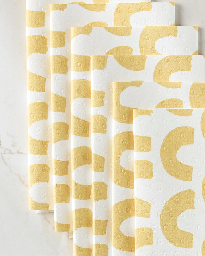 Five eco-friendly GEOMETRY "Sunny Hills" paper towels with a bold yellow and white abstract pattern—curved yellow shapes and small raised Gs—are arranged in a fan on a marble surface.