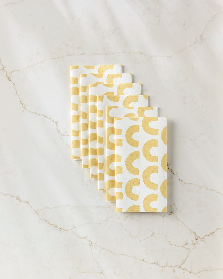 Seven stacked GEOMETRY Sunny Hills white reusable paper towels with a yellow abstract semicircle pattern, arranged diagonally on light beige marble.
