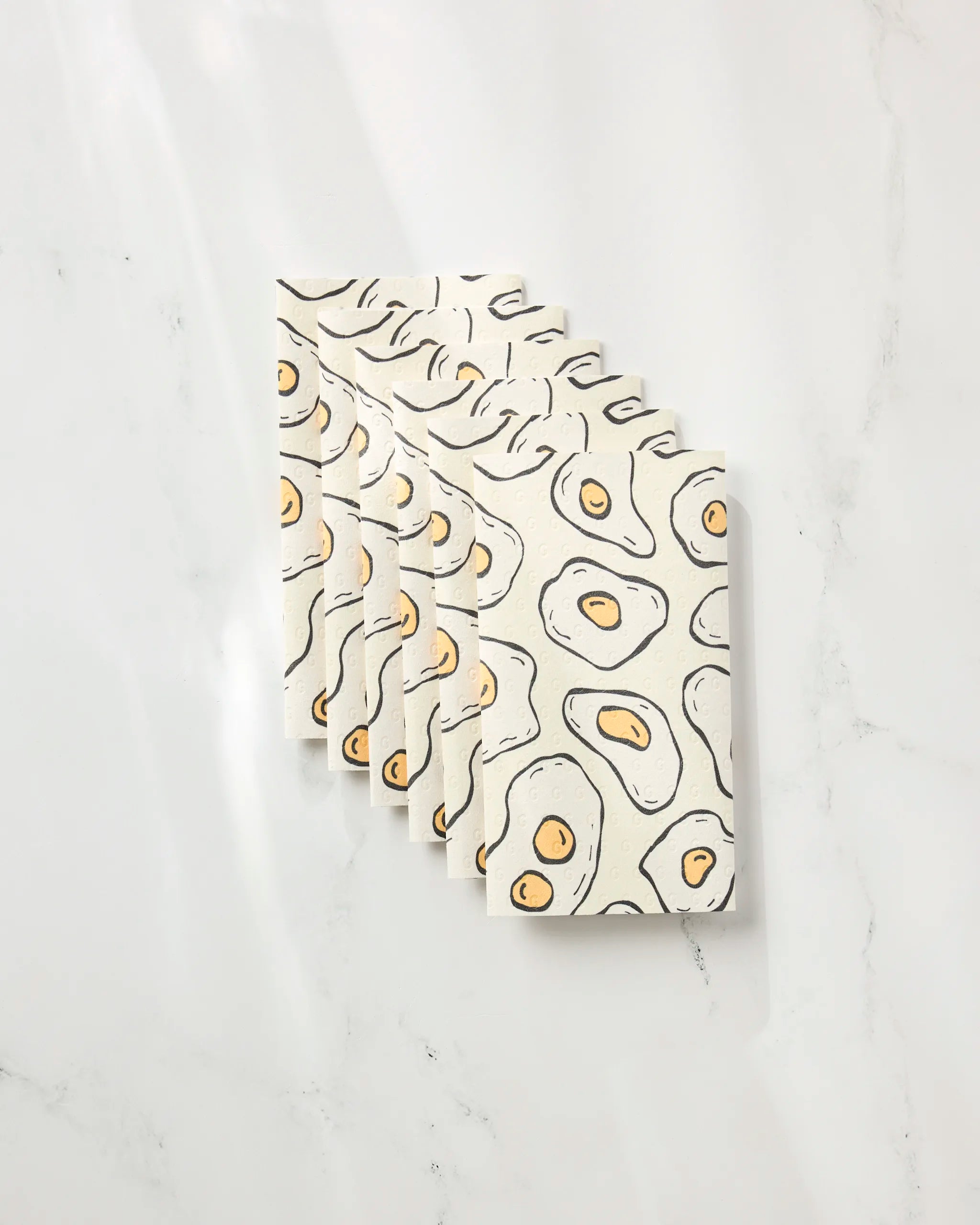 Six "Sunny Side Eggs" notebooks by Rosa Kichline, featuring an abstract fried egg pattern with black outlines and yellow yolks on a cream cover, are stacked diagonally on a white marble-like surface.