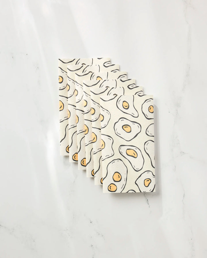 Six "Sunny Side Eggs" notebooks by Rosa Kichline, featuring an abstract fried egg pattern with black outlines and yellow yolks on a cream cover, are stacked diagonally on a white marble-like surface.