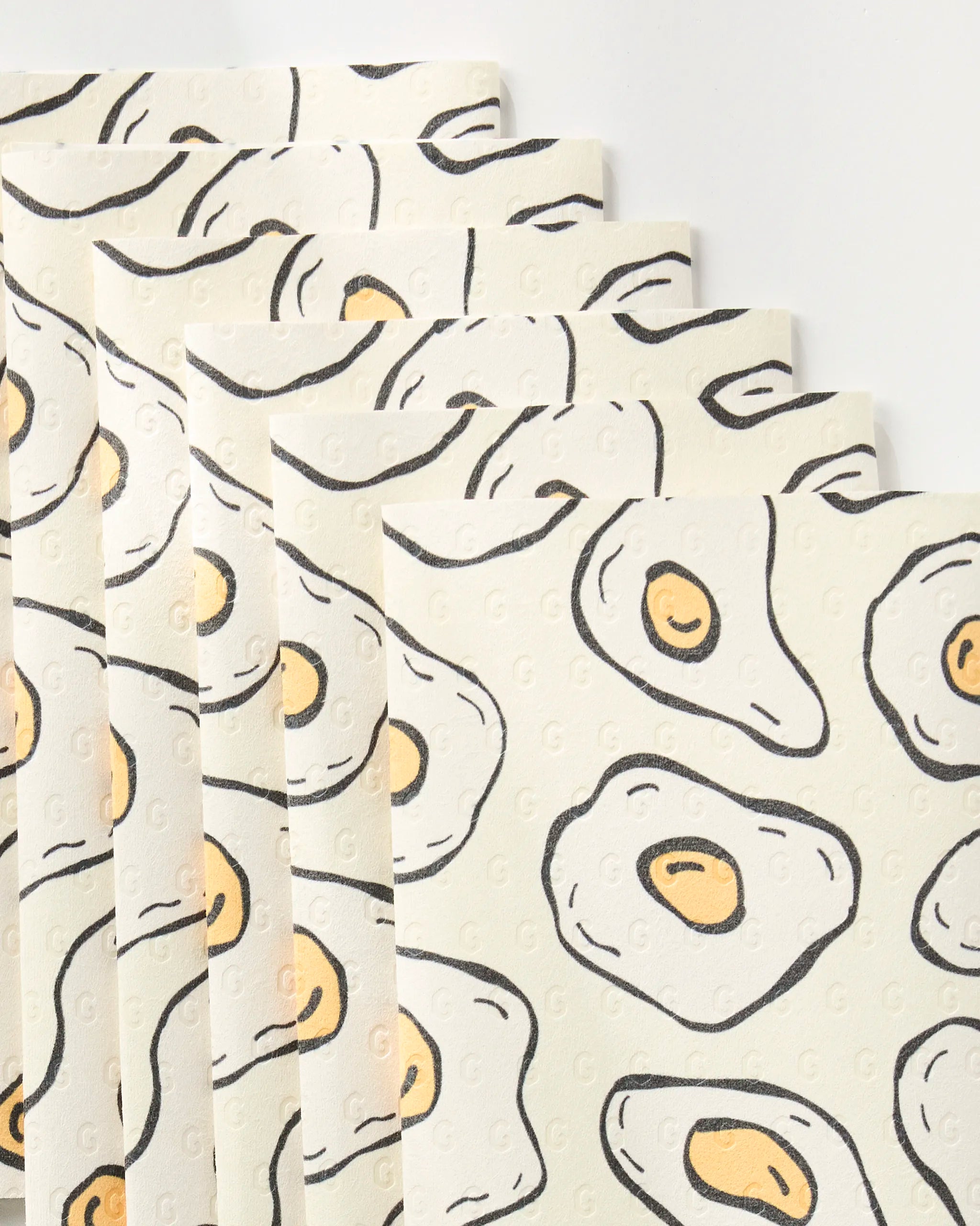Six "Sunny Side Eggs" paper towels by Rosa Kichline, eco-friendly sheets featuring a repeating cartoon fried egg pattern with yellow yolks and white edges, displayed fanned out on a white background.