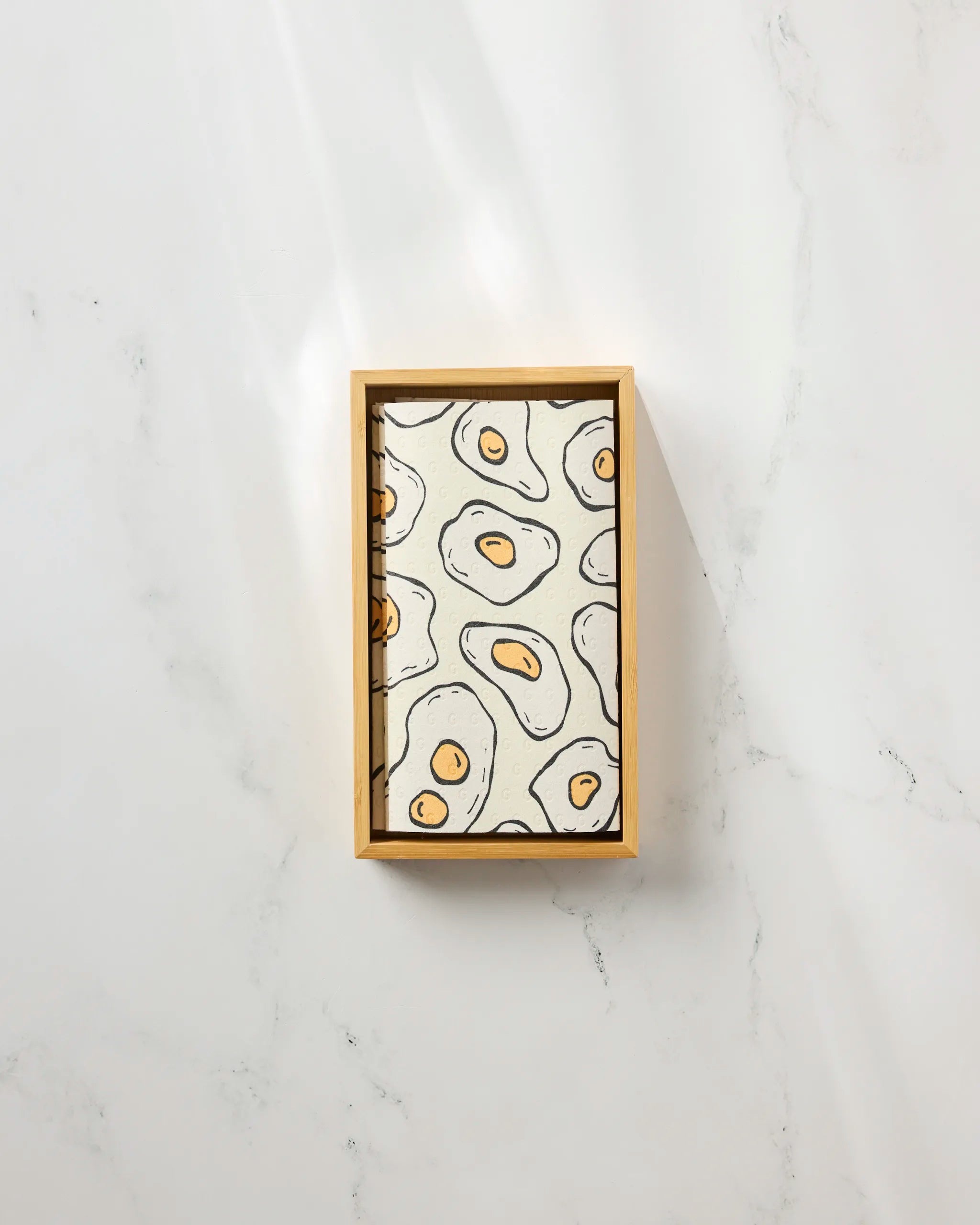 The "Sunny Side Eggs" rectangular wooden tray by Rosa Kichline features a cartoon fried egg pattern and is paired with eco-friendly paper towels, all displayed on a white marble surface.
