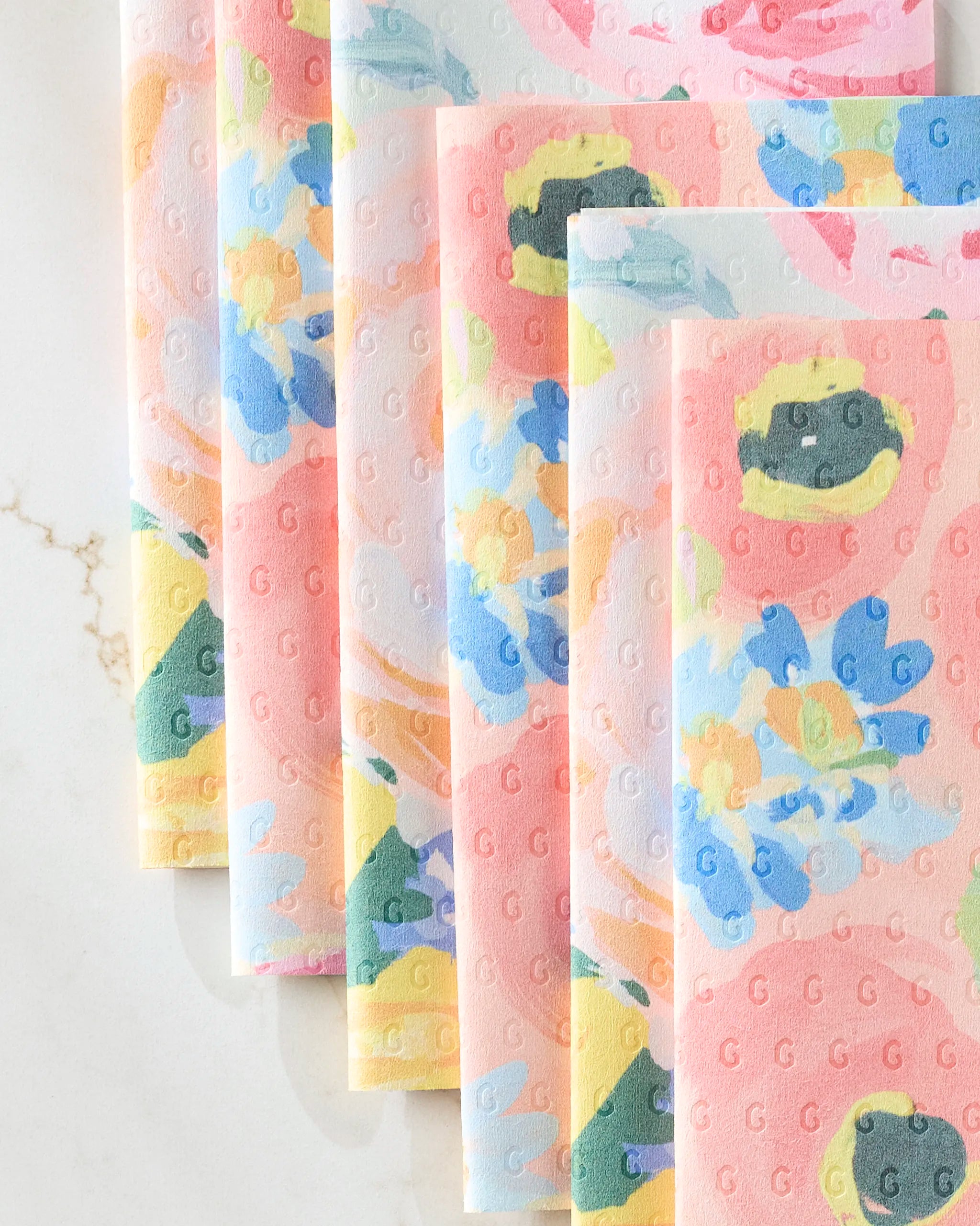Five eco-friendly Suzzy and Margot paper napkins by Ania Zwara Art, featuring pastel floral patterns in pink, yellow, blue, and green, are neatly fanned out on a light marble surface.