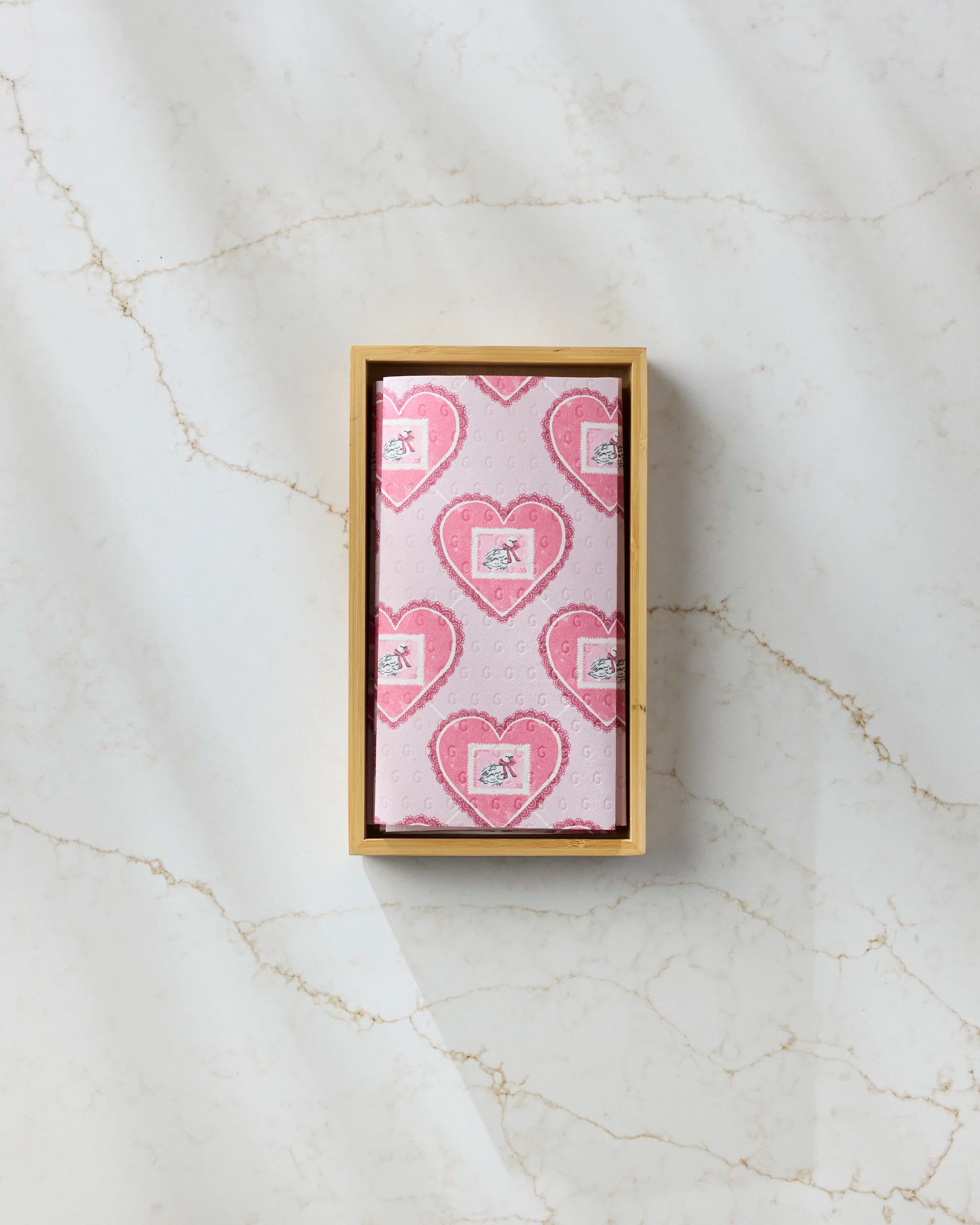 A rectangular box of Geometry's Swan Argyle eco-friendly kitchen towels, wrapped in heart-patterned paper, rests on a white marble surface with light brown veins.