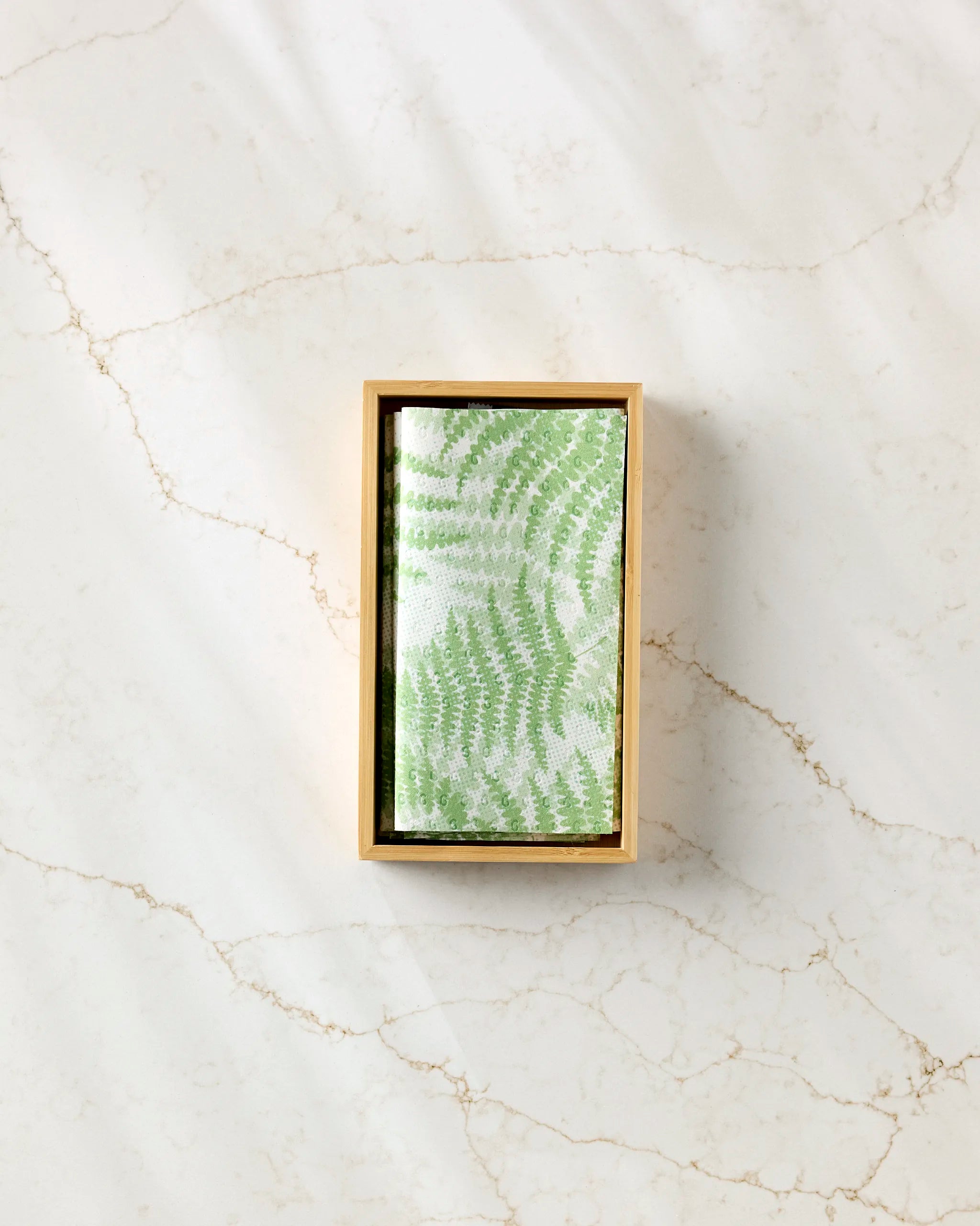 The GEOMETRY Swaying Leaf rectangular wooden box holds a folded white cloth featuring a green leaf pattern, resembling eco-friendly kitchen towels, displayed on white marble with beige veining.