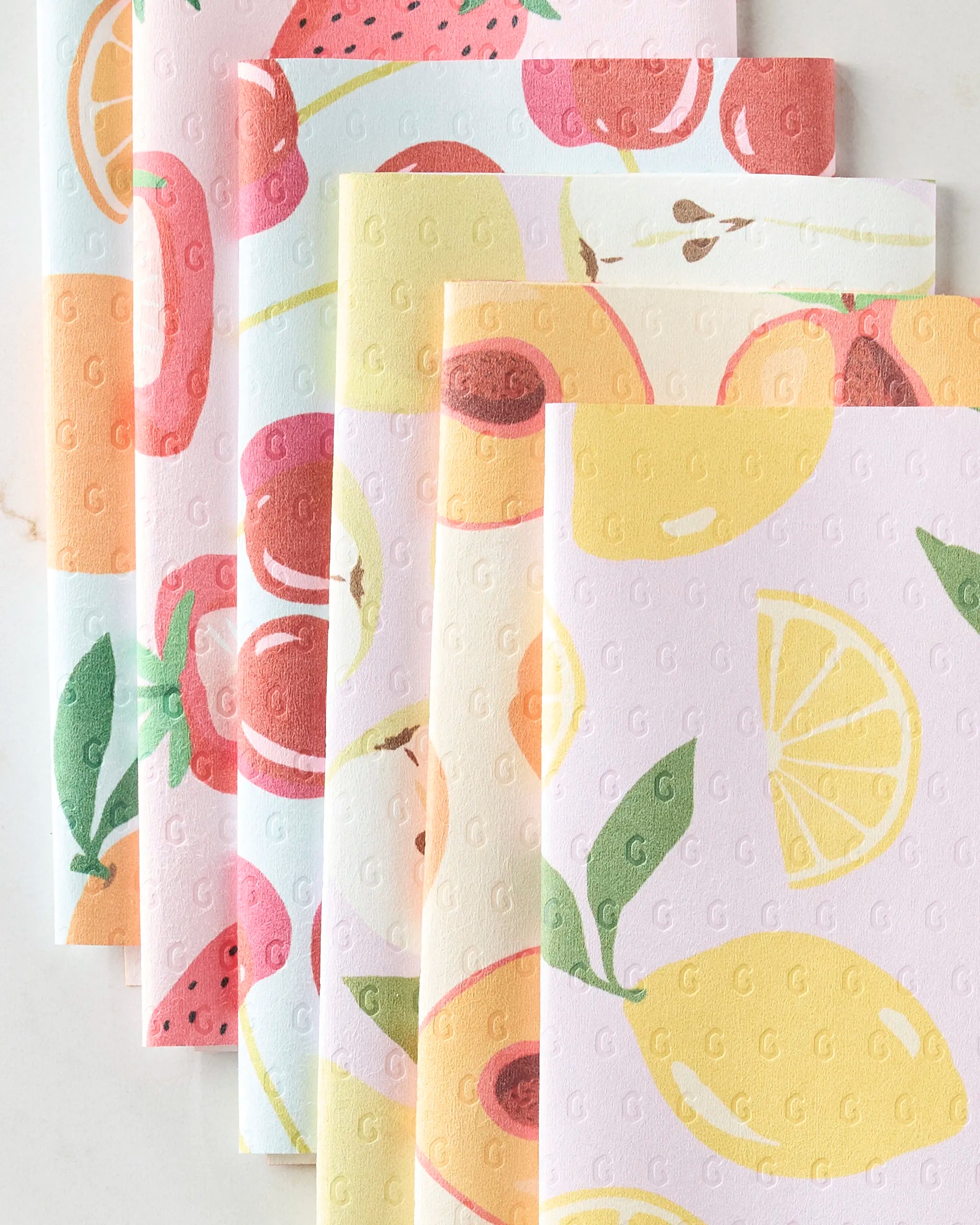 Five reusable GEOMETRY Sweet Summer Mix 1 paper towels, each with a unique fruit pattern—watermelon, apples, peaches, and lemons—fan out on a light surface. The eco-friendly cloths display a subtle embossed G design.
