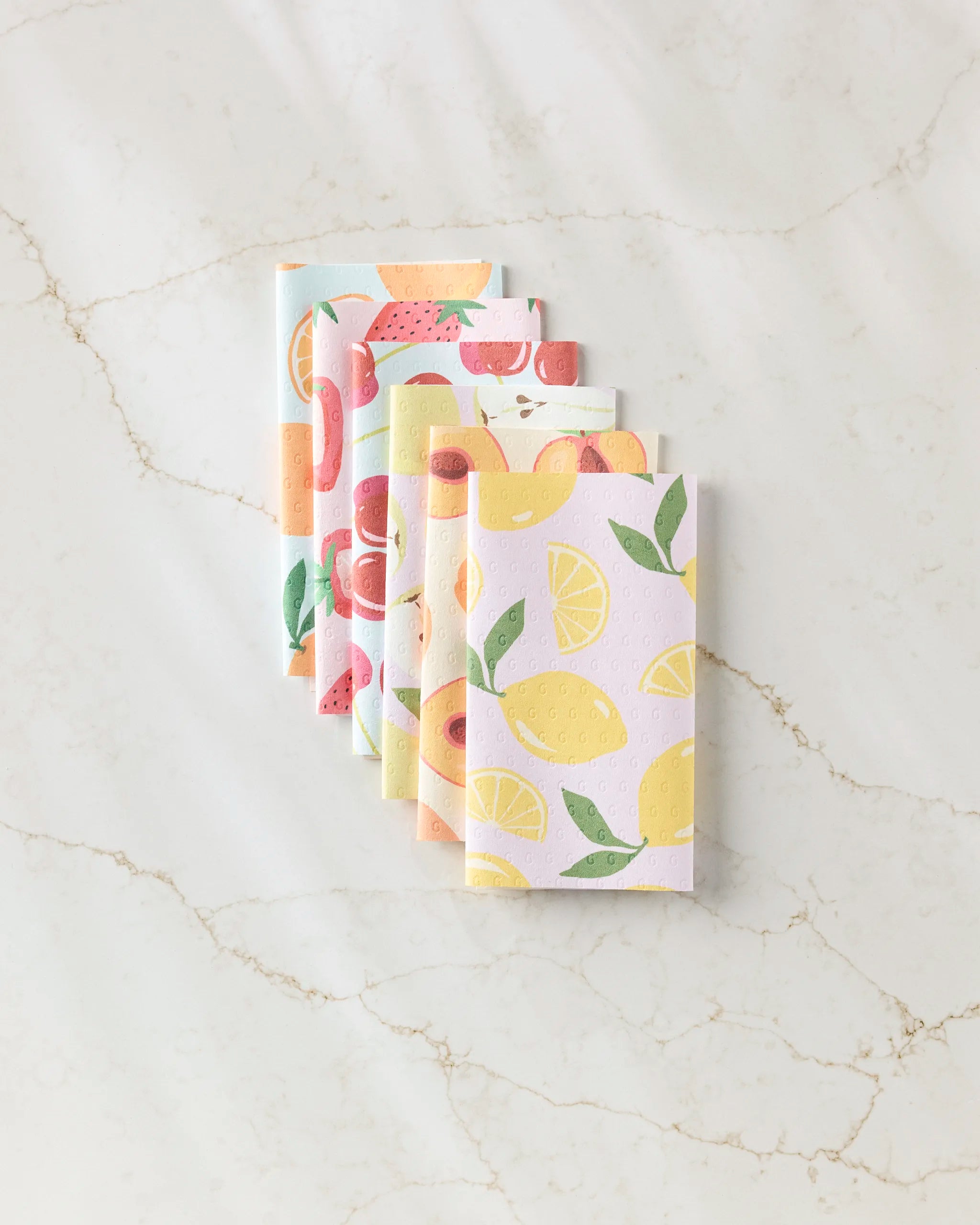 GEOMETRY’s Sweet Summer Mix 1 features five folded reusable paper towels with bright fruit patterns, arranged in a fan shape on a light marble surface for an eco-friendly touch.