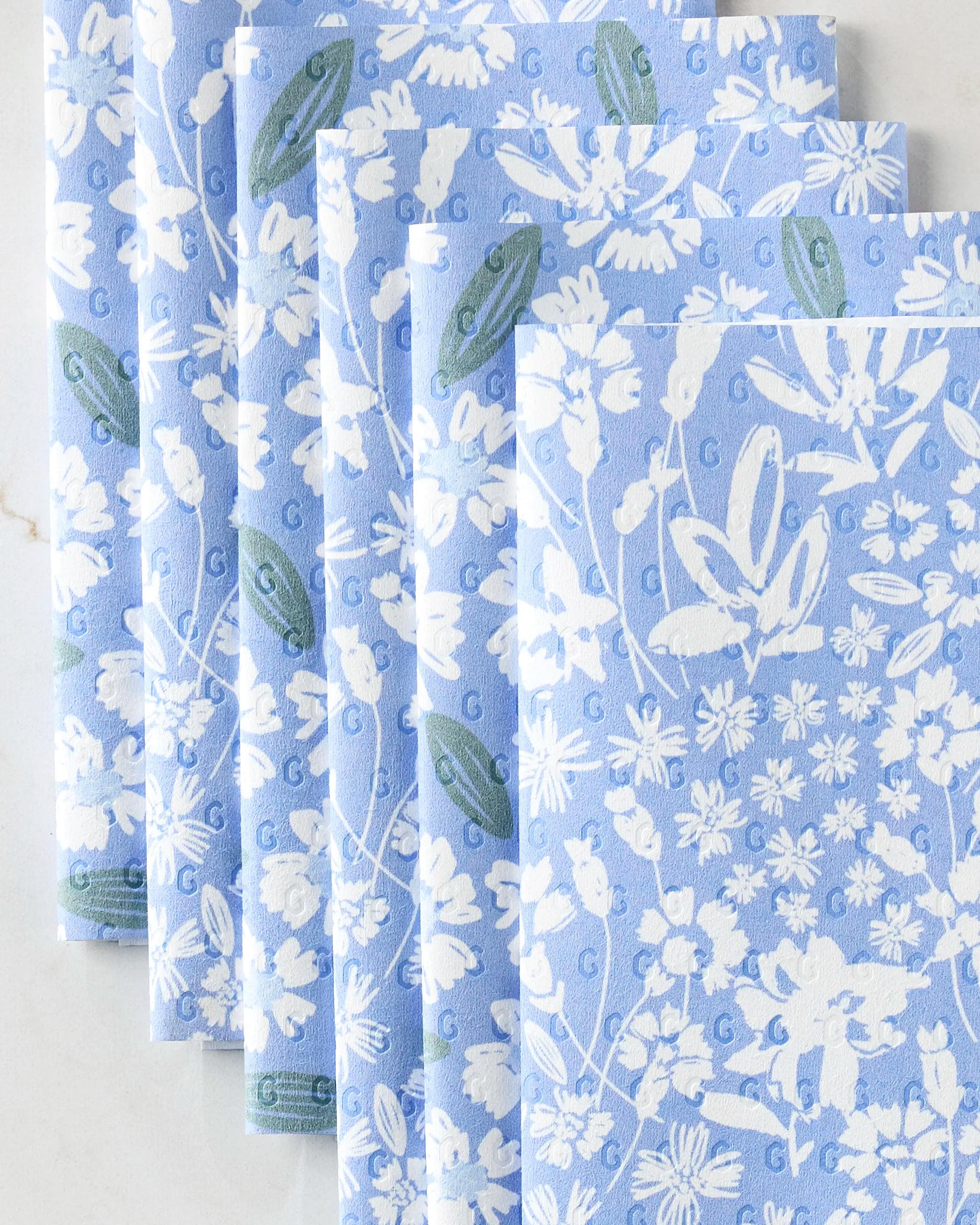 Five folded Sweetie Floral reusable paper towels by Hufton Studio, featuring a blue background with white floral patterns and green leaf accents, are arranged in a neat, overlapping row on a light surface.