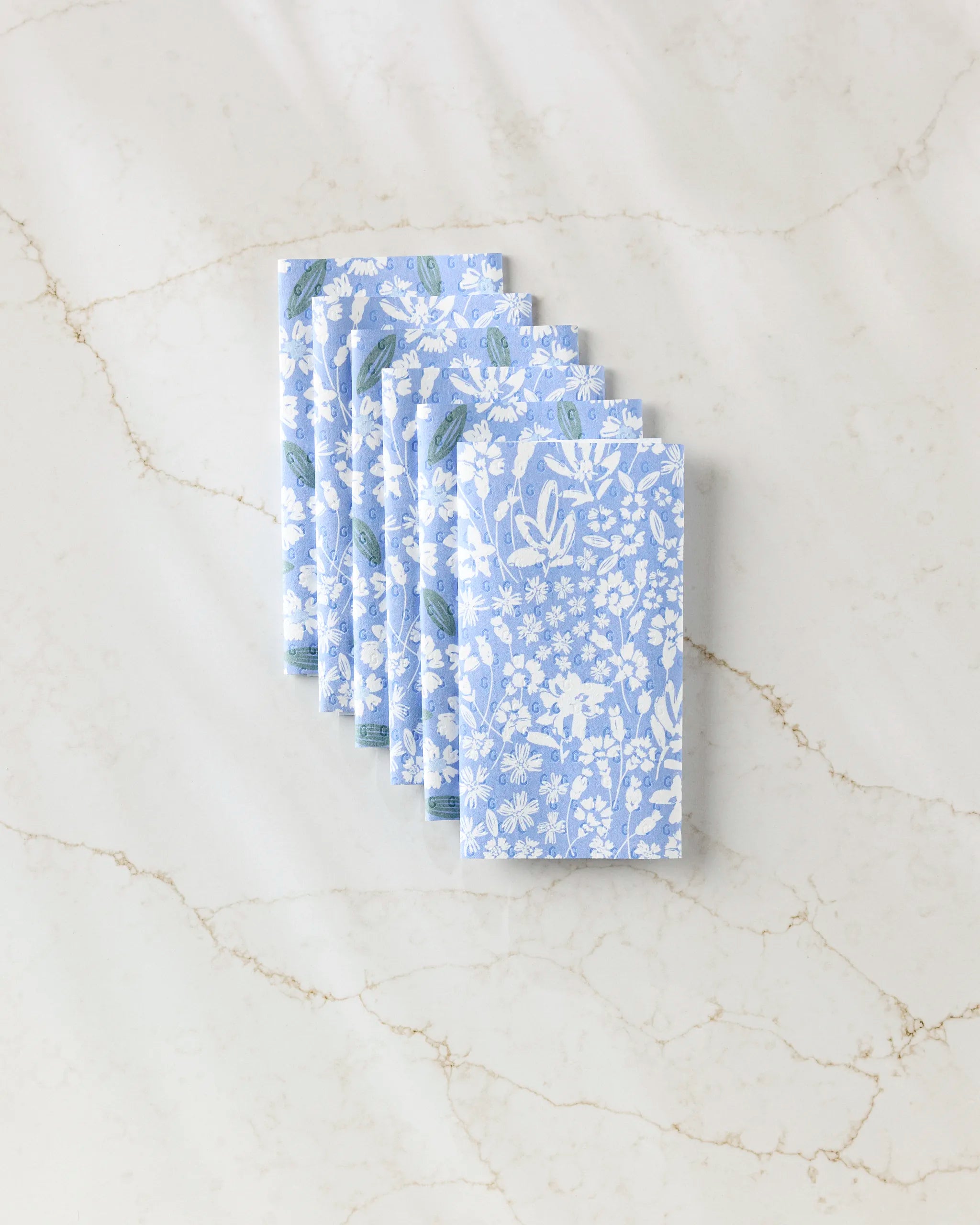 Six neatly folded Sweetie Floral blue napkins by Hufton Studio, featuring a white floral pattern and displayed on white marble—ideal as eco-friendly kitchen towels for any sustainable home.