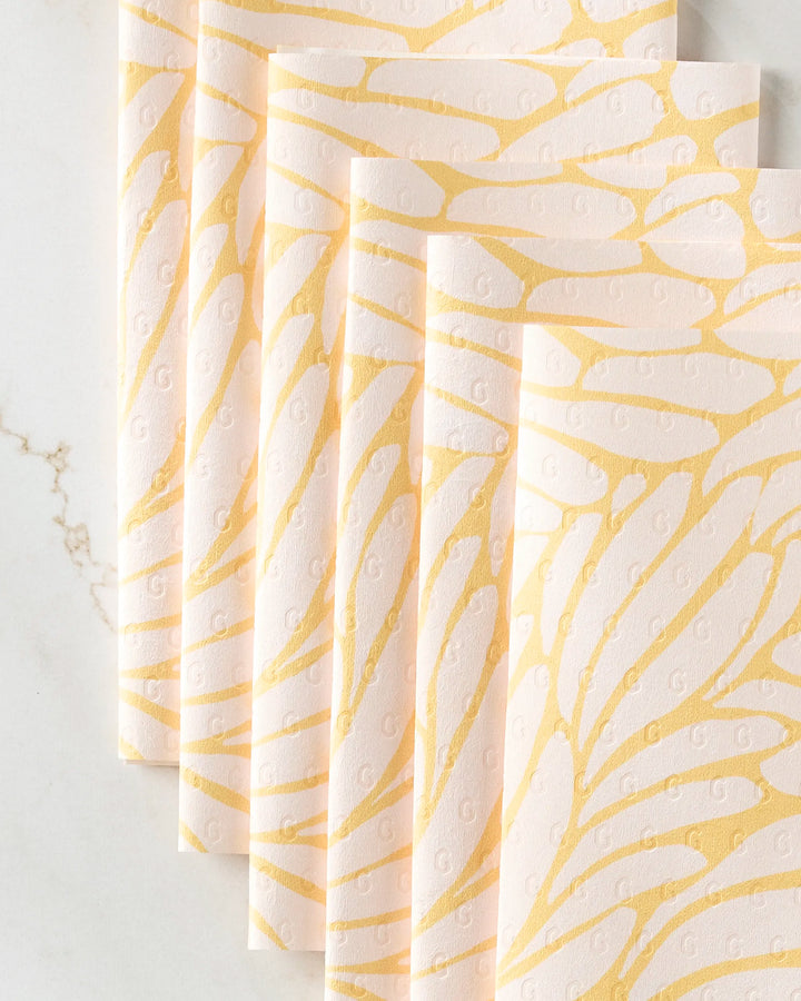 Five pale pink "Tenderhearted" reusable paper towels by GEOMETRY, featuring a gold abstract wing pattern, are arranged in an overlapping fan display on a white marble surface.