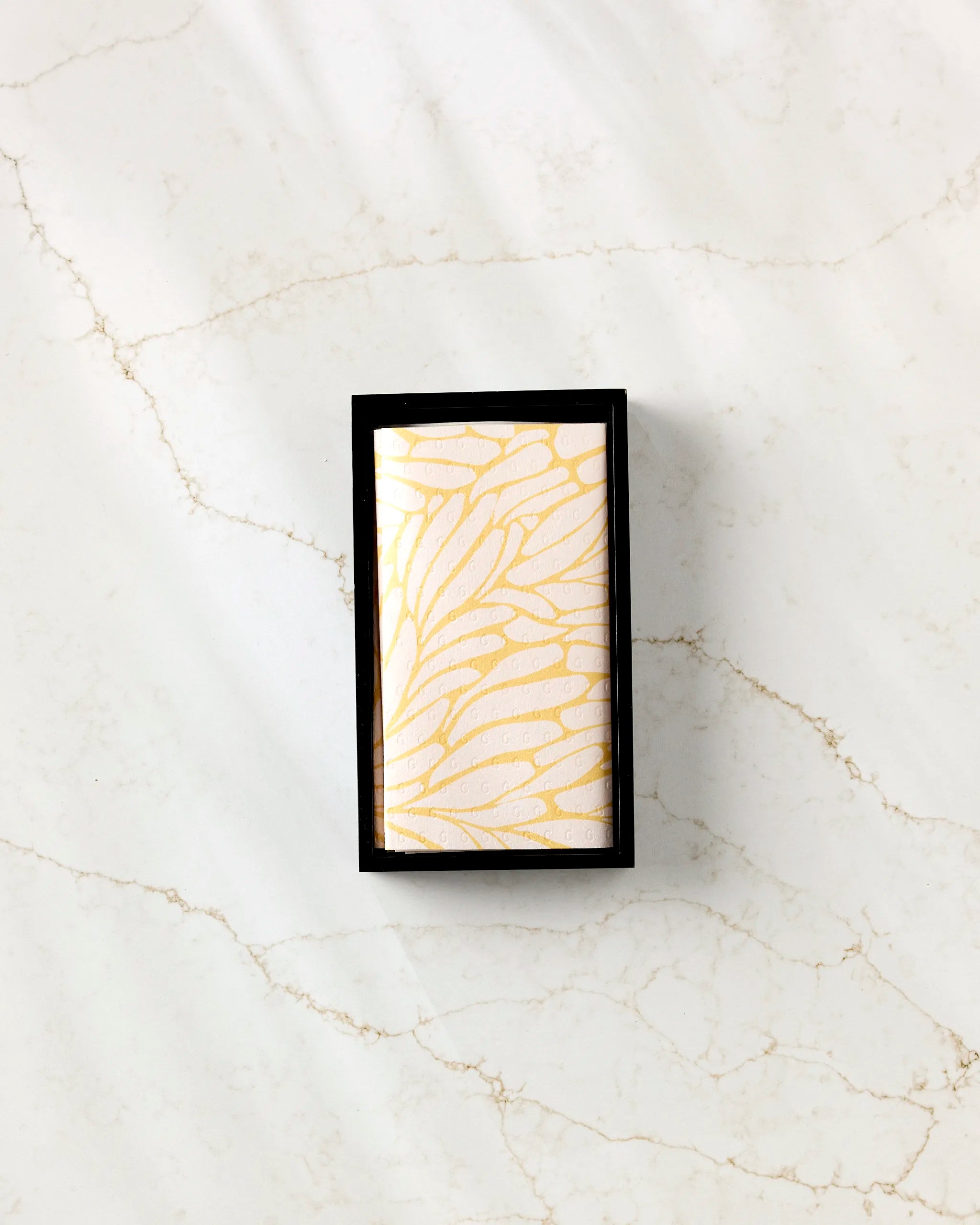 The GEOMETRY Tenderhearted rectangular soap, featuring a gold feather-like pattern, is displayed in a black box on white marble with light brown veining beside folded eco-friendly kitchen towels.