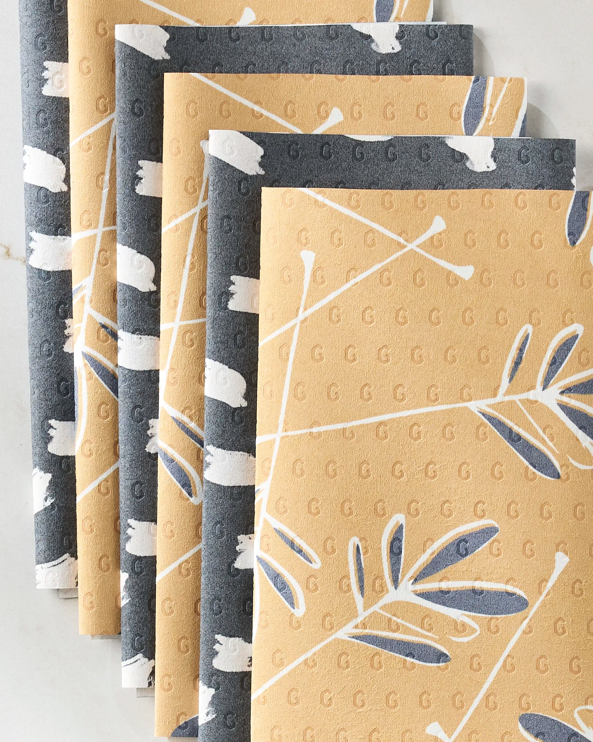 Five folded, reusable paper towels from GEOMETRY’s Tess And Dark collection feature a textured surface with white, blue, and gray leafy branches and embossed Gs on a light background. Eco-friendly with a modern yellow and gray floral design.