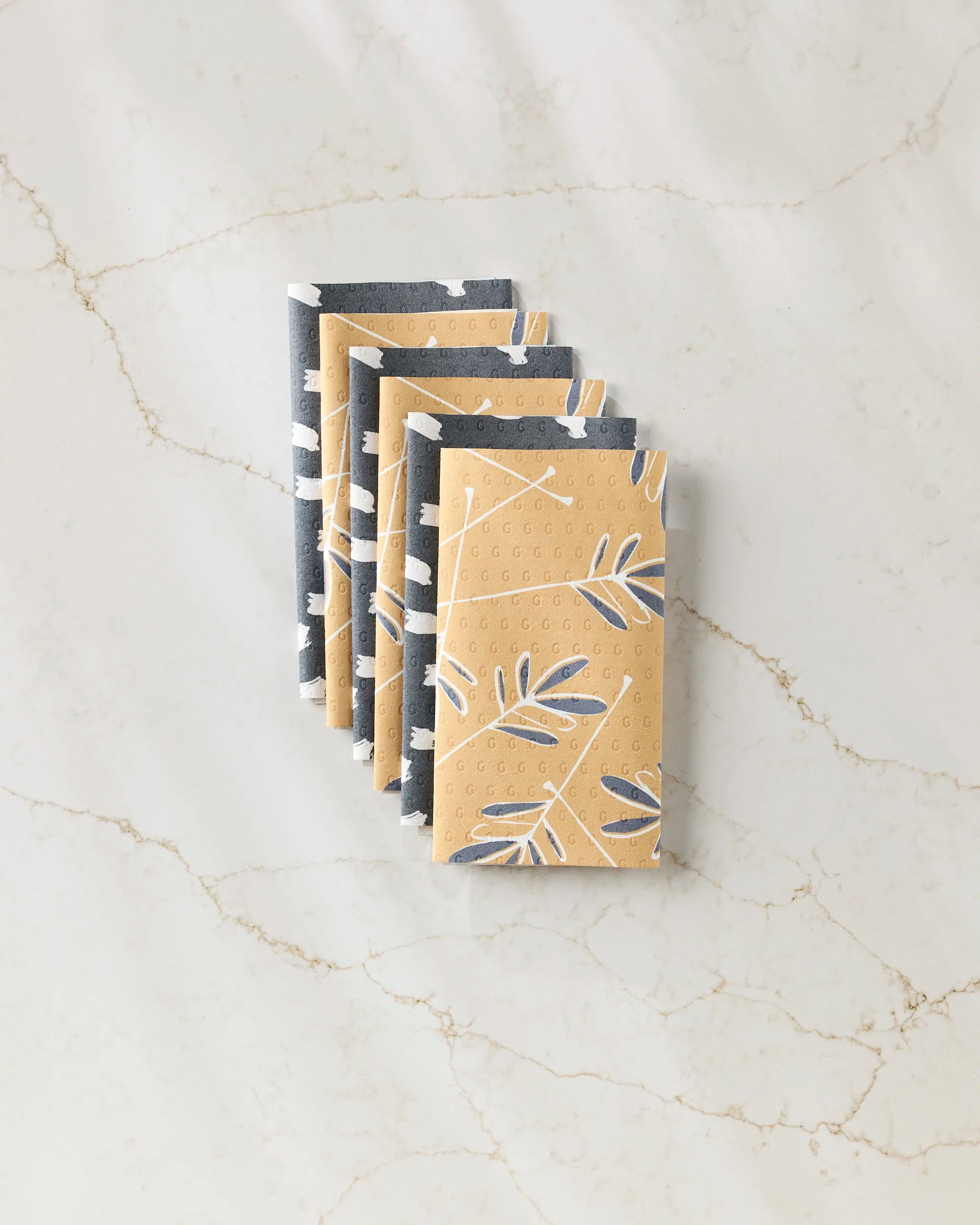Four folded Tess And Dark reusable paper towels by GEOMETRY, featuring a beige and gray leaf pattern, are neatly stacked on a white marble surface with light brown veins.
