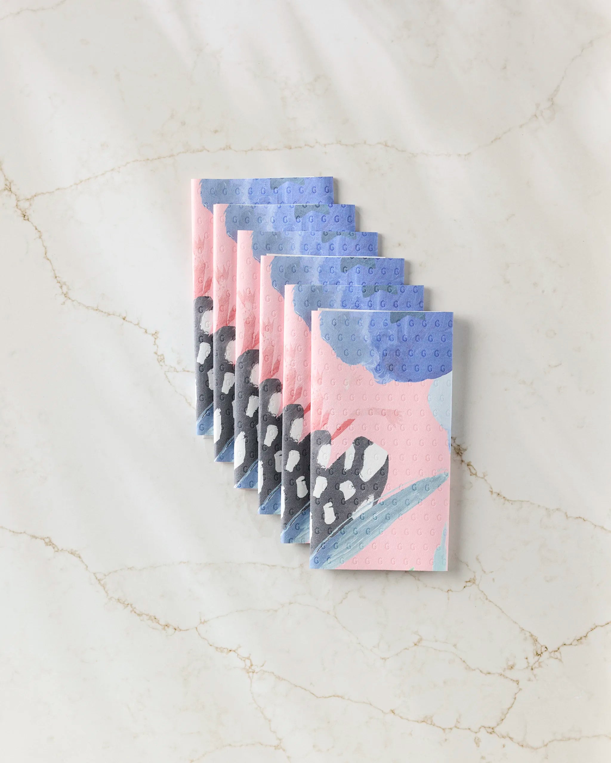 Seven folded GEOMETRY "Thinking Of You" reusable paper towels, featuring an abstract pink, blue, black, and white design, are neatly arranged on a white marble surface with light brown veins.