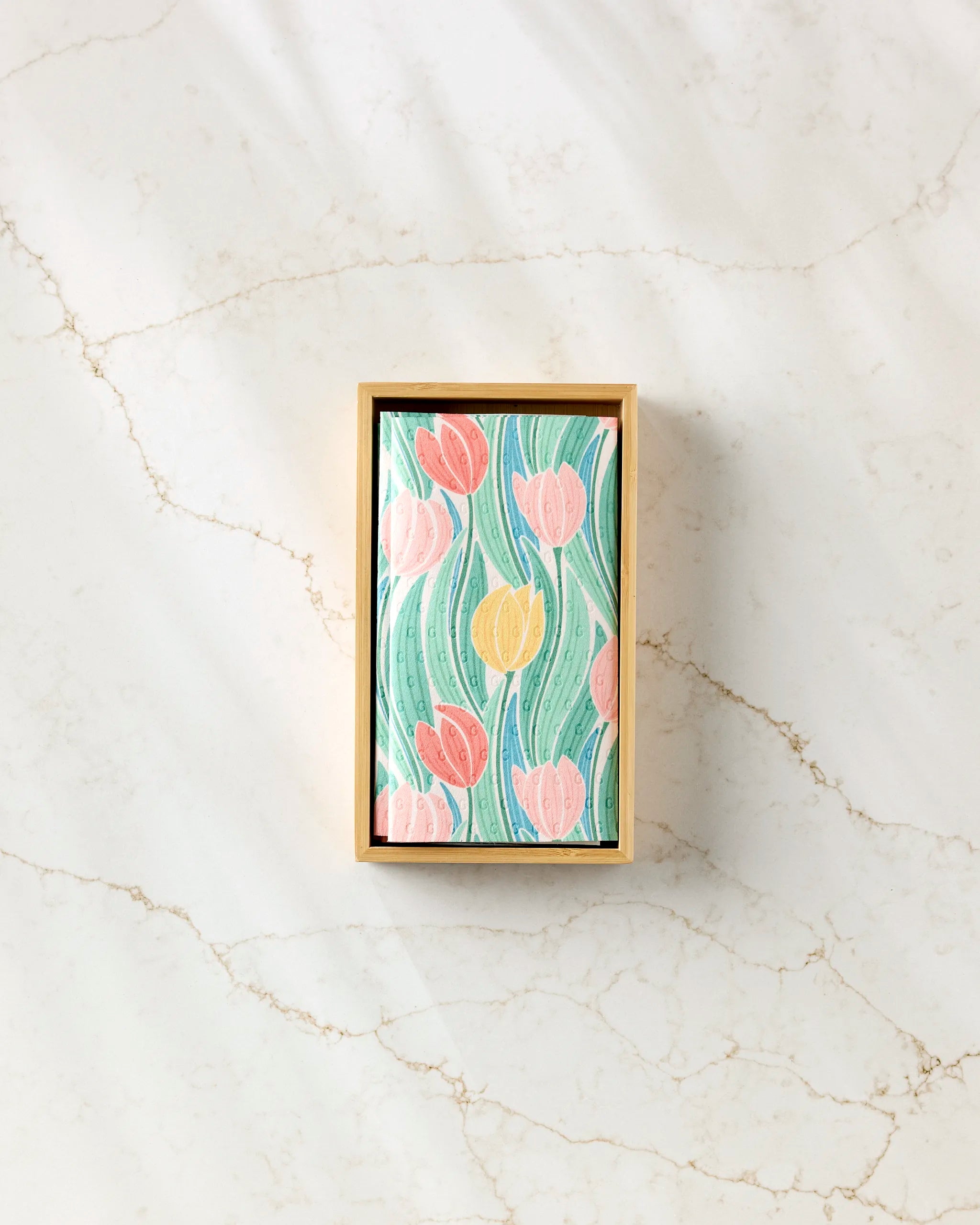 The Tranquil Tulips rectangular box by Sarah Daugherty, with a floral lid of pink and yellow tulips and green leaves, rests on a light marble surface and is designed to hold eco-friendly paper towels.