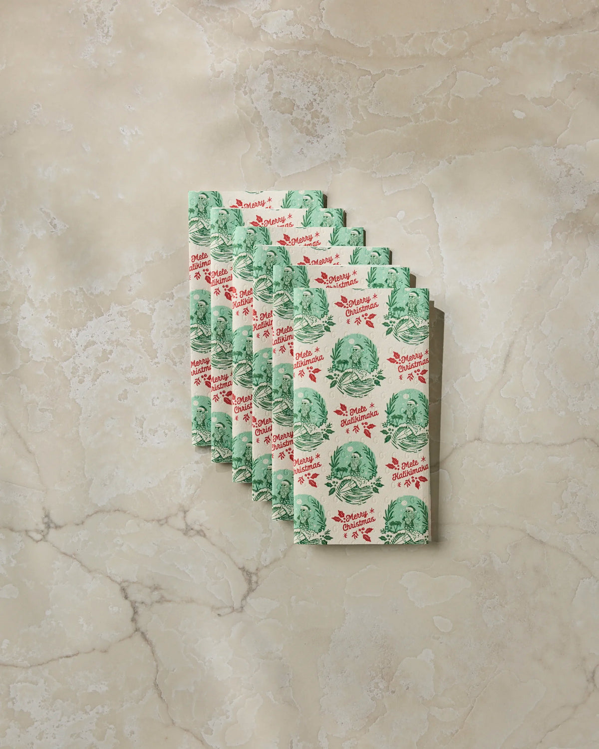 Eight folded, eco-friendly Geometry “Tropical Tidings” paper napkins featuring green cabbage illustrations and red cursive text are neatly arranged in a vintage style on a marble surface. The napkins are stacked in a slightly fanned pattern.