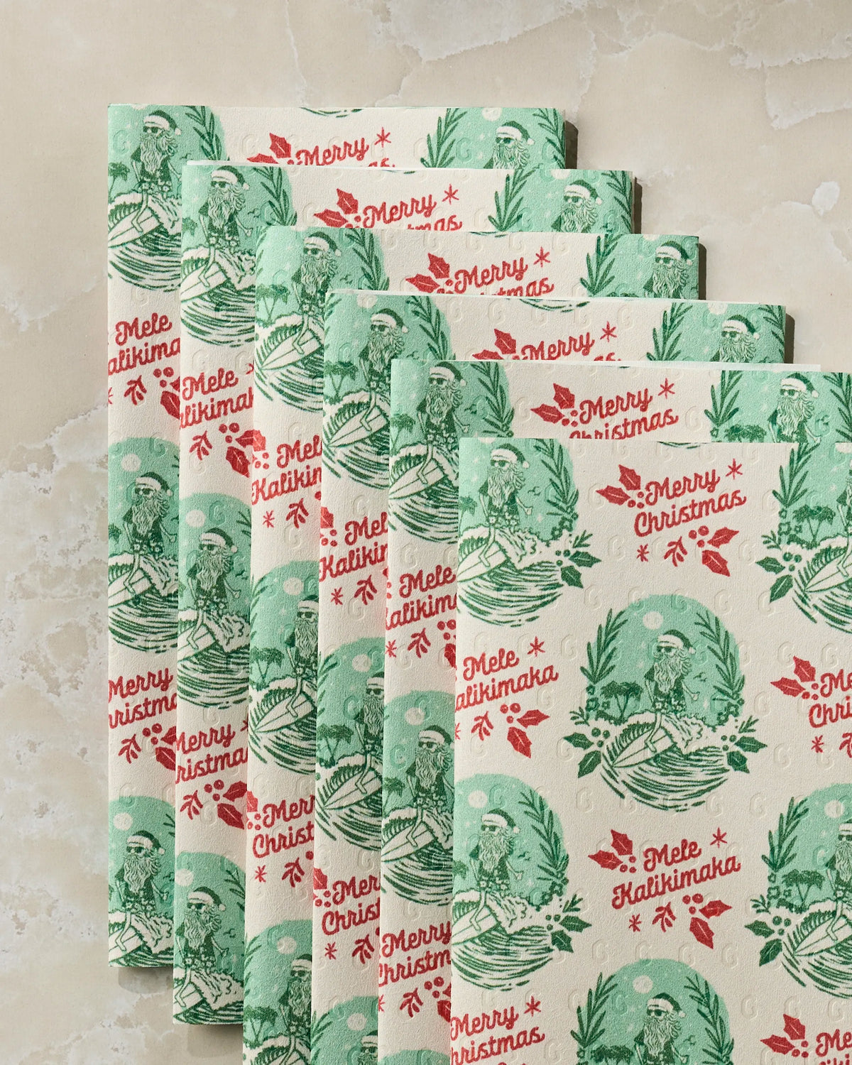 Set of six festive “Tropical Tidings” napkins by Geometry, made from eco-friendly paper towels, with green and red “Merry Christmas” and “Mele Kalikimaka” lettering and cheerful illustrations of a person with palm trees and holiday decorations, all beautifully displayed on a light marble surface.