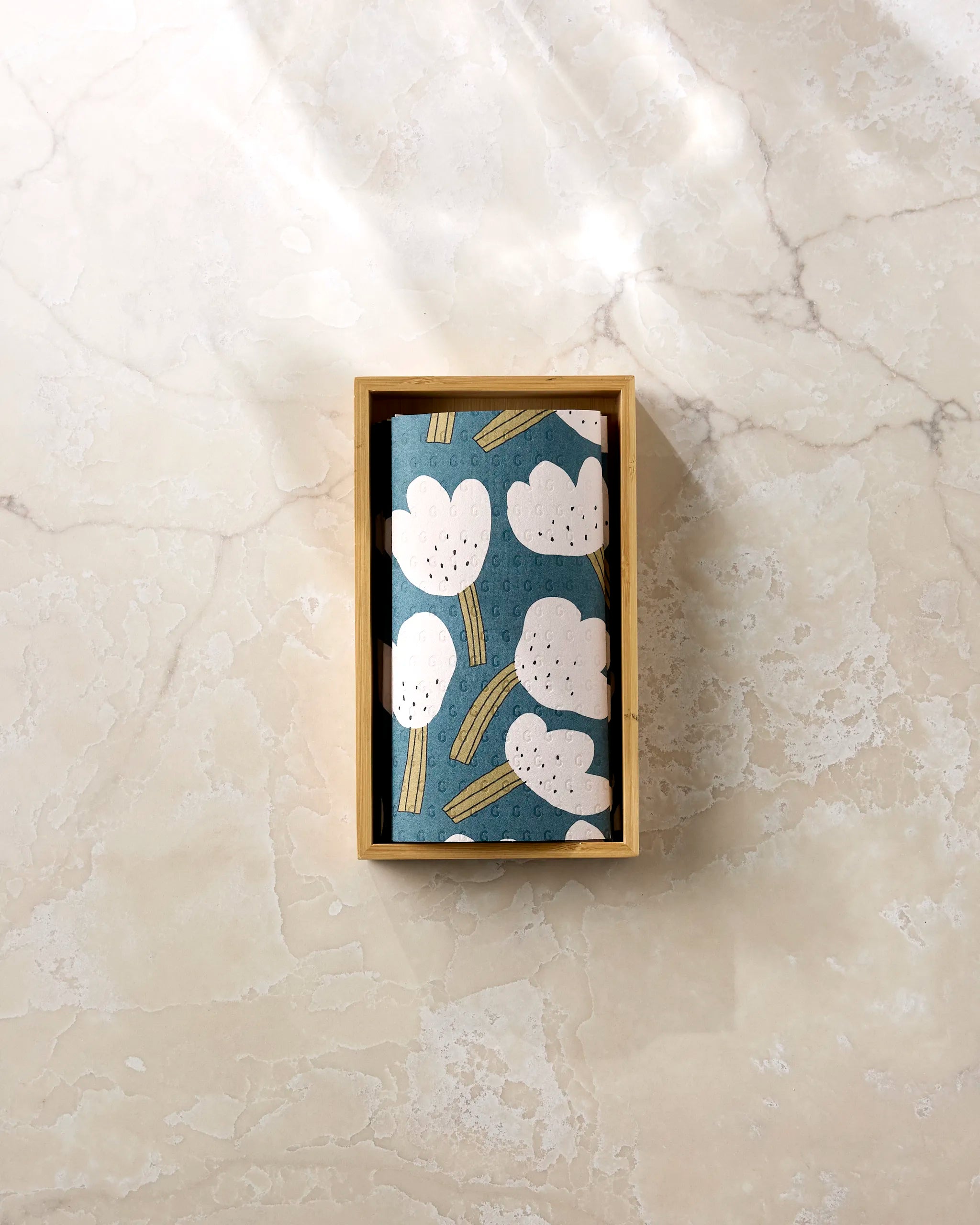 A small wooden box with Jennifer Bouron's "Tulips" notebook—featuring white abstract flowers and yellow stems on blue—placed on a light marble surface, paired with eco-friendly paper towels for a sustainable touch.