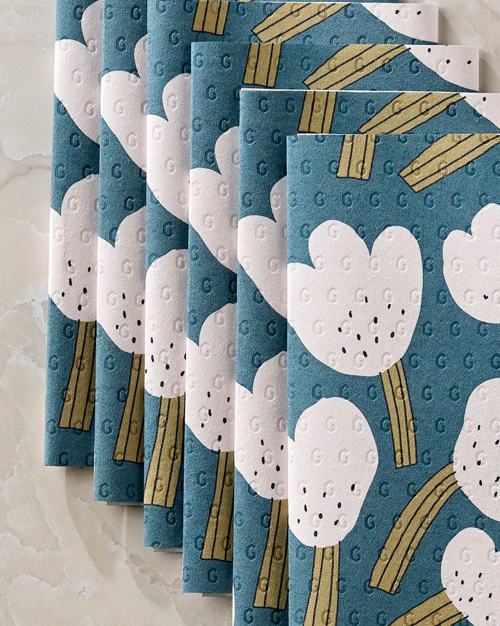 Six folded "Tulips" napkins by Jennifer Bouron, featuring a playful blue design with white florals and yellow stems, are neatly overlapped in a row on a light marbled surface.