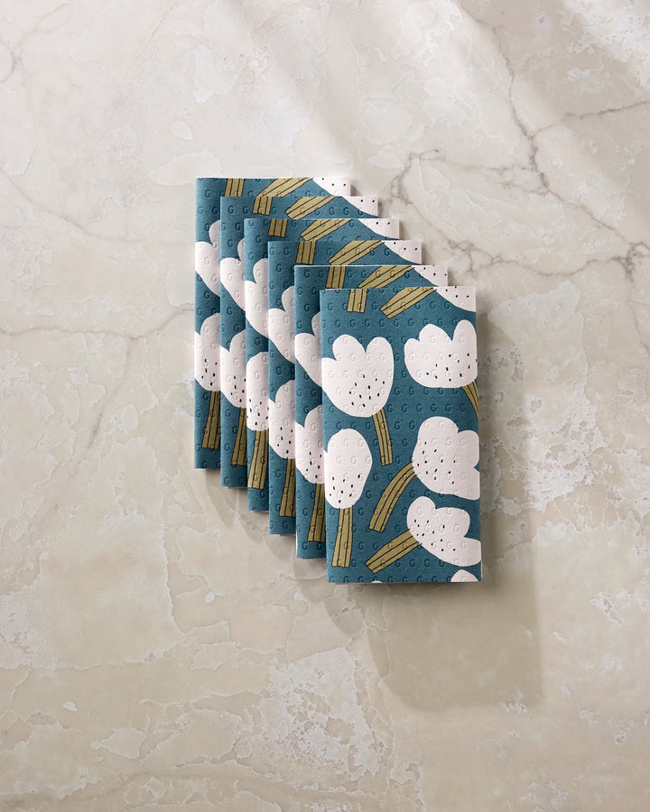 Six neatly folded Tulips reusable paper towels by Jennifer Bouron, featuring blue fabric with white clouds and yellow lines, are arranged in a fan shape on a light marble surface.