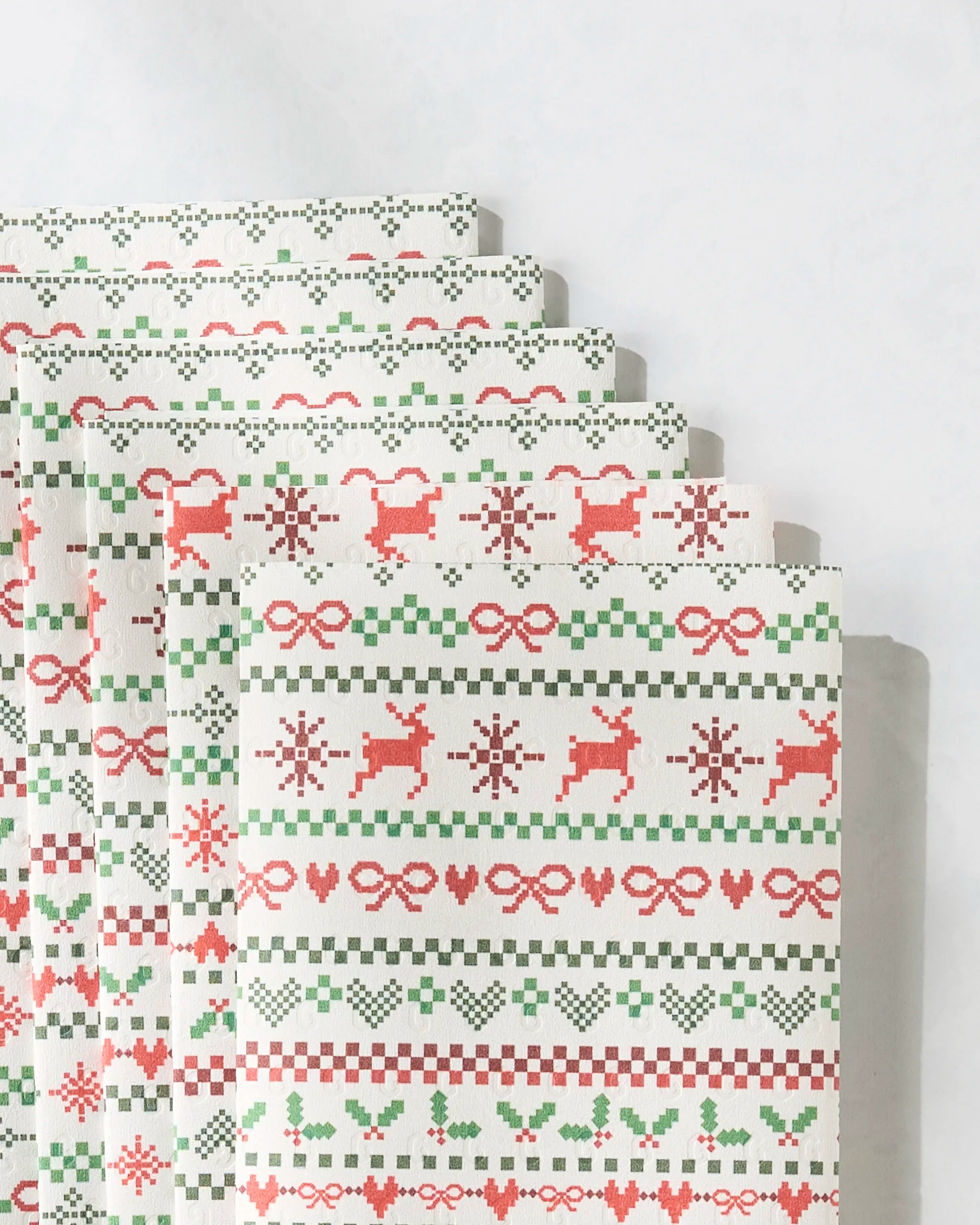 A neatly arranged stack of Geometry’s "Ugly Sweater Day" festive, eco-friendly paper towels features red and green holiday patterns—reindeer, snowflakes, hearts, bows, holly, and checkered lines—on a light surface.