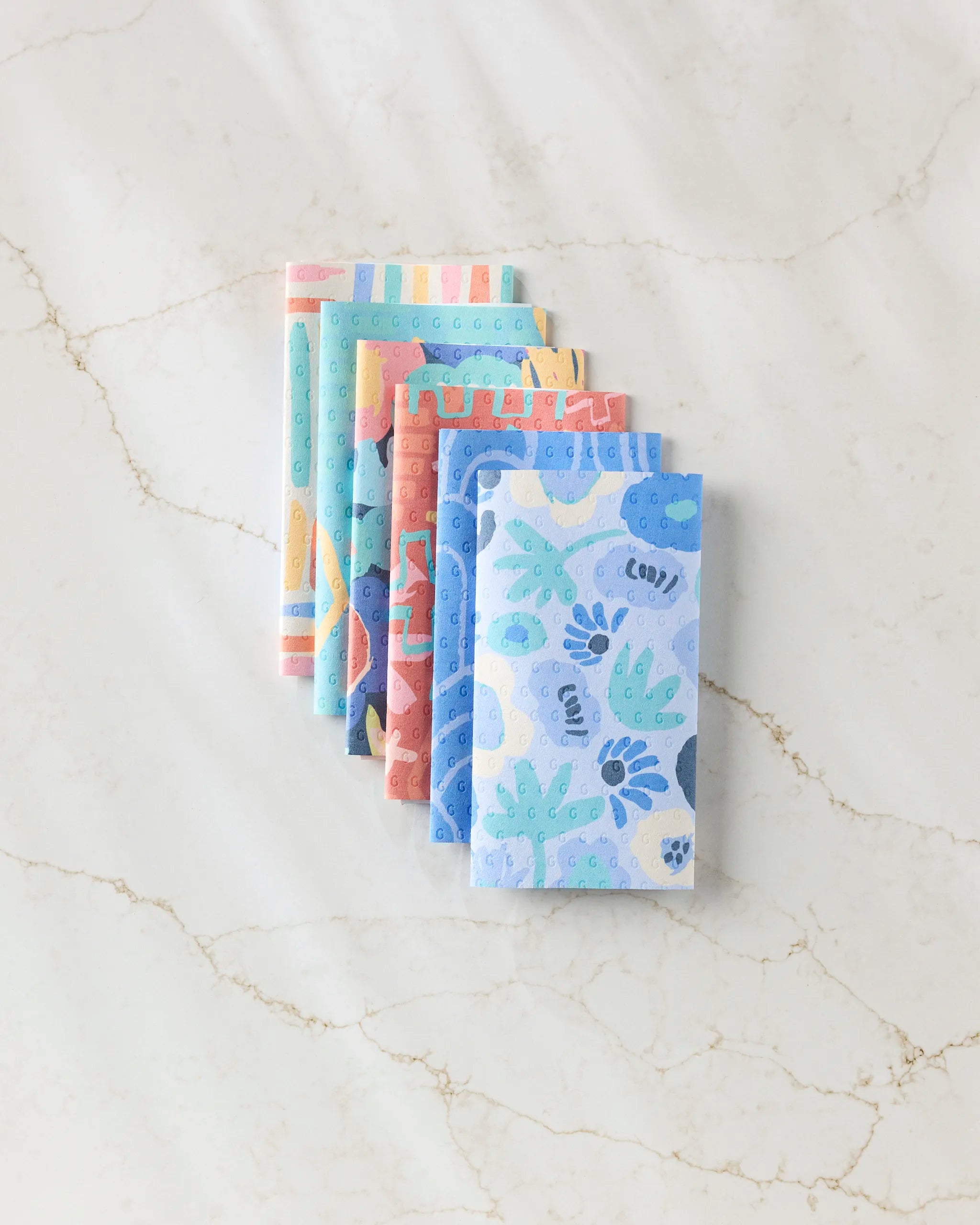 Five eco-friendly, reusable kitchen towels from Sareka Unique are stacked on a white marble surface. The Unique towels feature playful blue, green, and pink patterns—an ideal alternative to disposable paper towels.