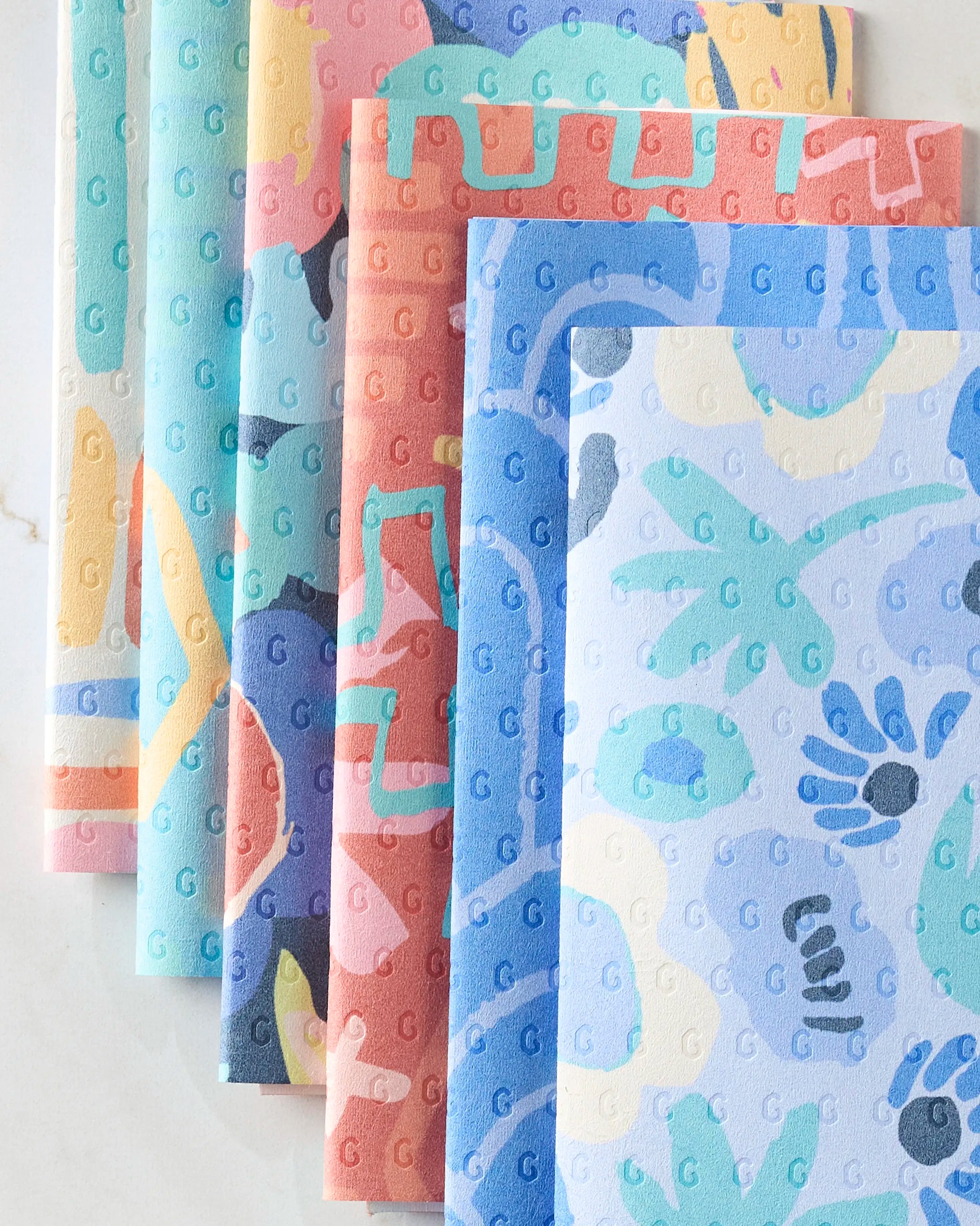 Unique by Sareka Unique presents colorful, patterned sheets with abstract shapes in pastel and vibrant tones. Featuring an embossed G pattern, these eco-friendly alternatives to paper towels are arranged on a light marble surface.