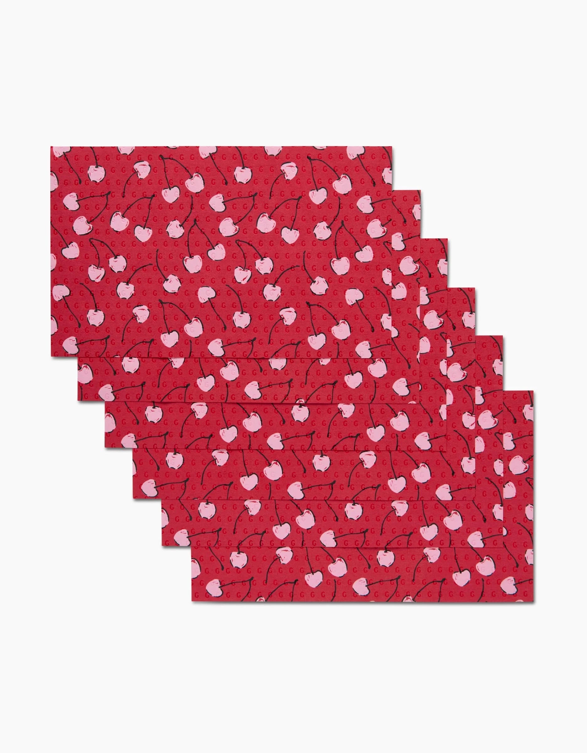 Six rectangular red placemats from Hufton Studio’s Valentines Fruit Cherry collection, featuring a white cherry pattern and made from eco-friendly kitchen towels, are stacked in a staggered arrangement against a white background.