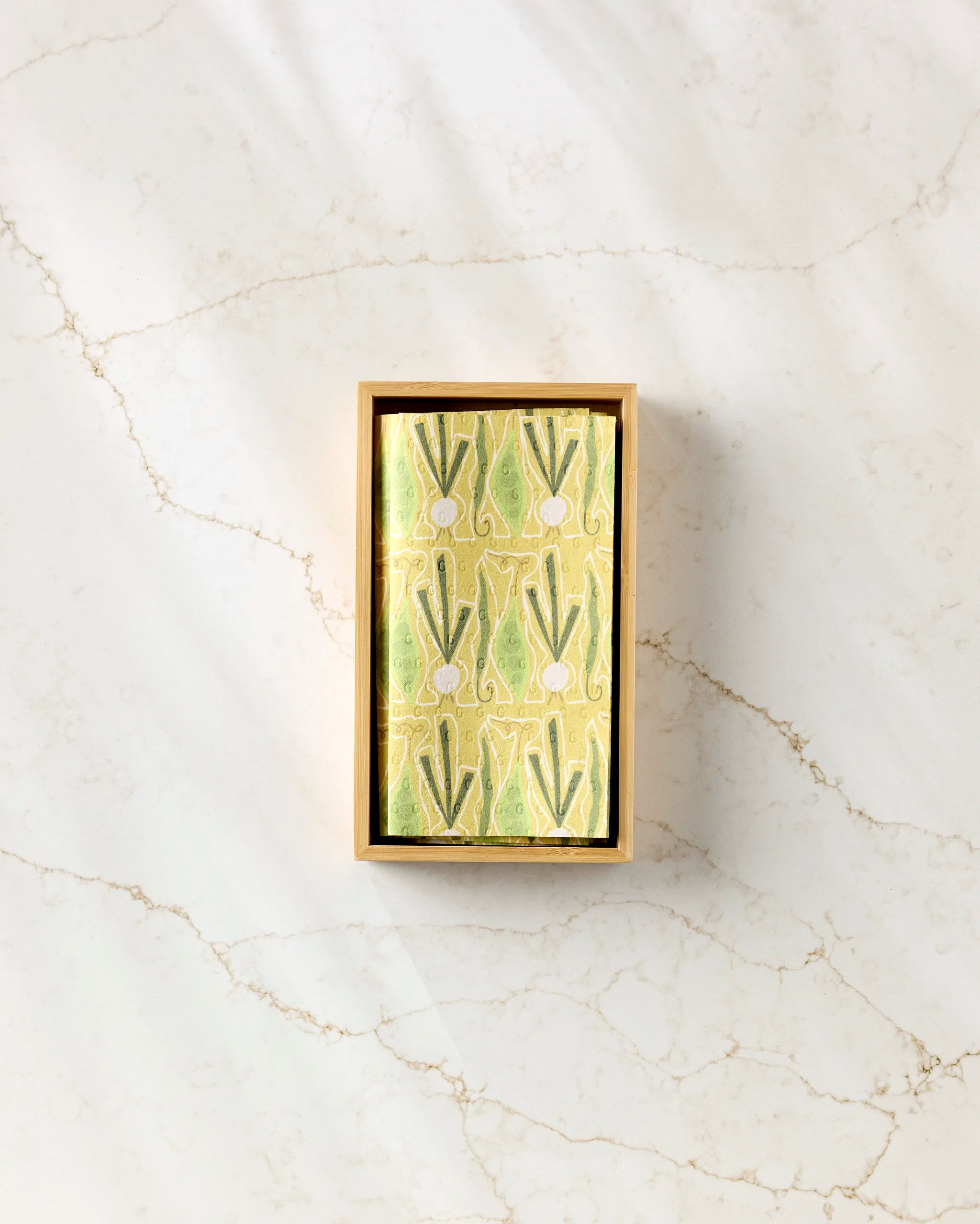 Veg on Parade by Lizzie Clark is a rectangular box with a decorative lid featuring green leaves and white circles on yellow, perfect for storing eco-friendly paper towels and displayed on a light marble surface.