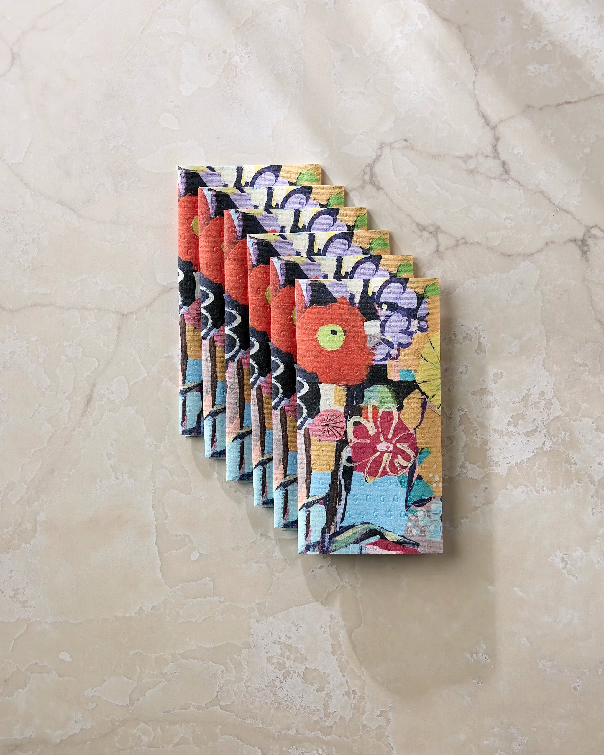Seven vibrant Vera reusable paper towels by Ania Zwara Art, each with red, yellow, and purple floral patterns, are neatly arranged in a diagonal row on a beige marble surface.