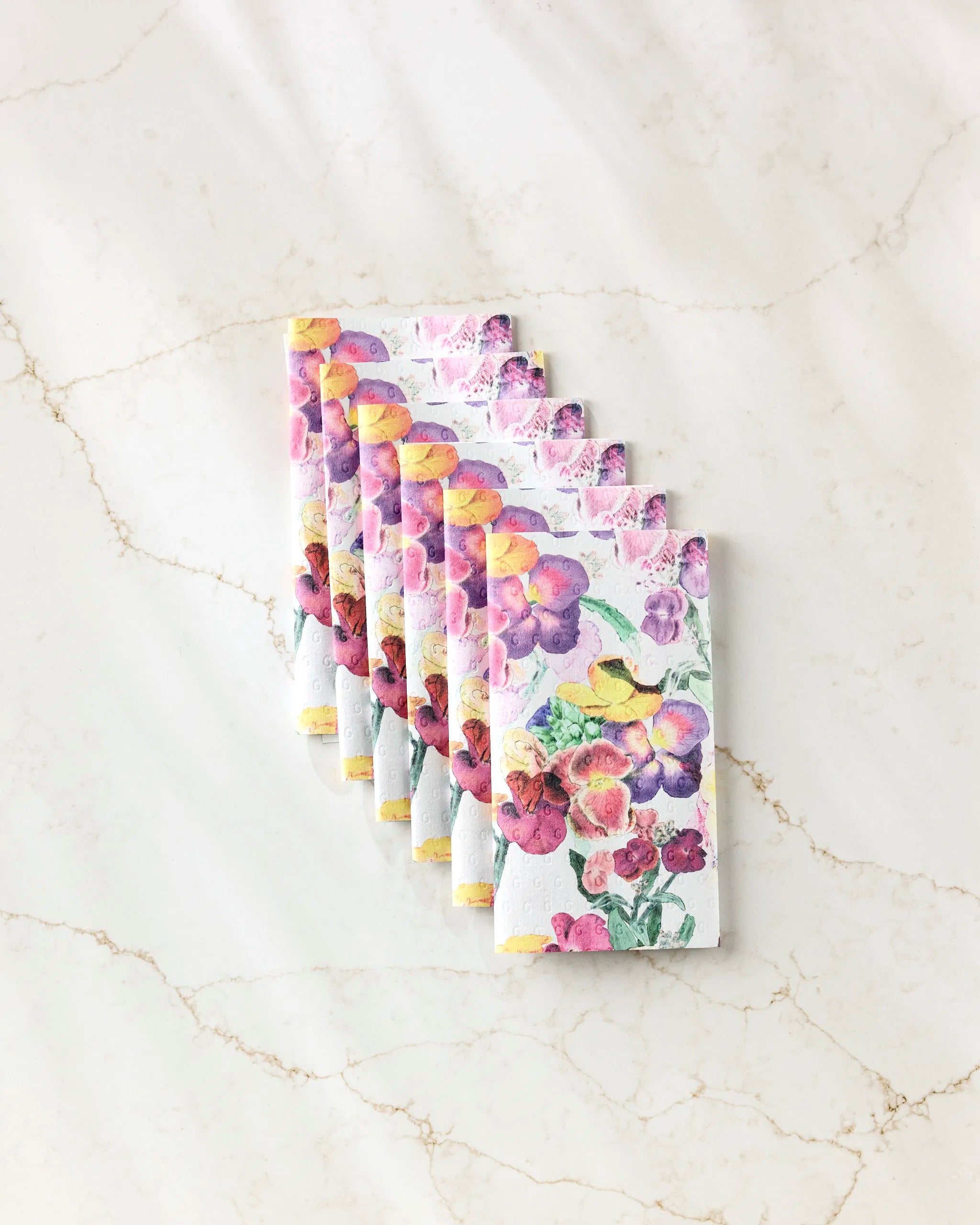 Seven Wallflower chocolate bars by Pascale Dilger, featuring bright floral watercolor packaging, are arranged in a neat diagonal line on a light marble surface, evoking the freshness of reusable paper towels.
