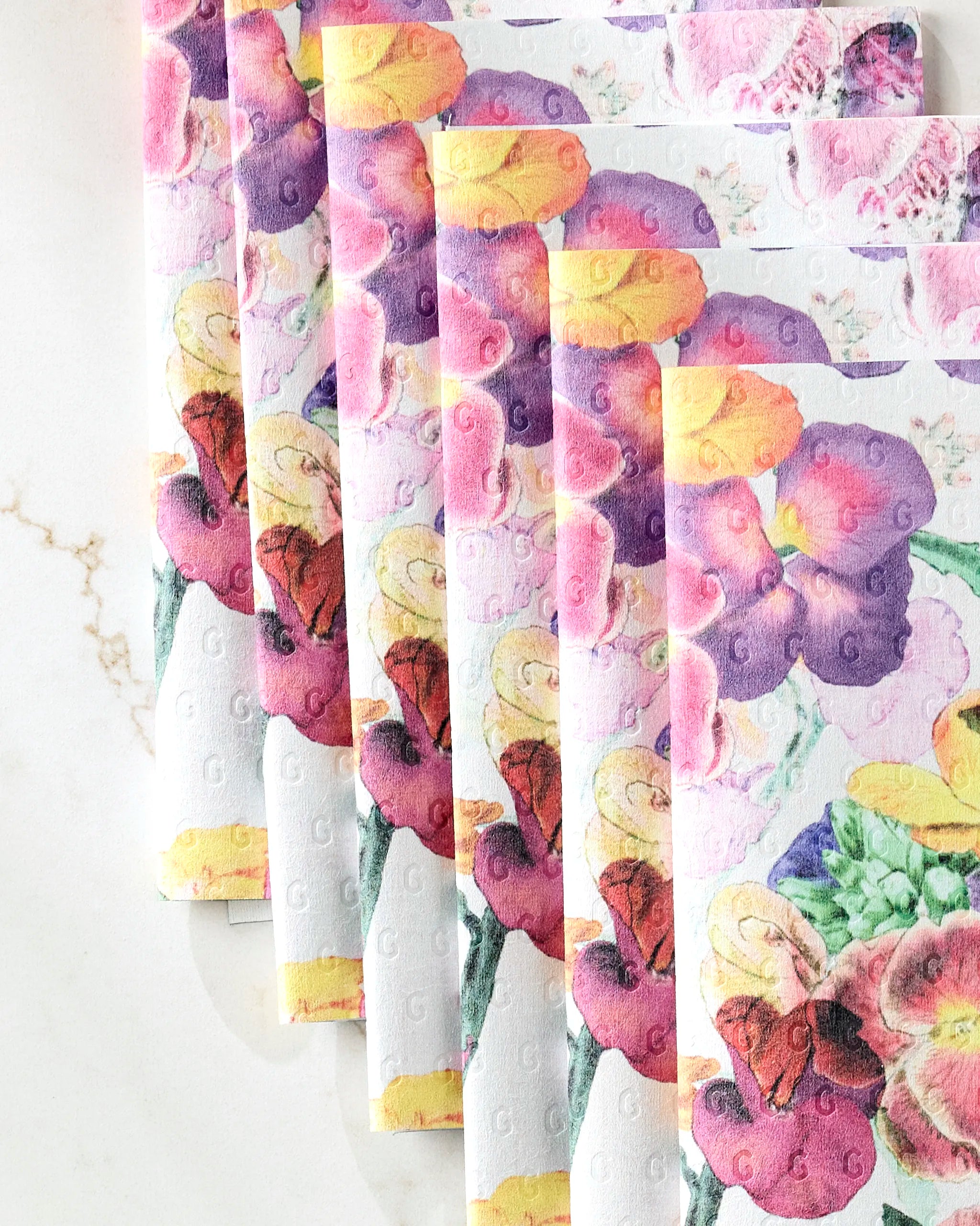 A stack of Wallflower reusable paper towels by Pascale Dilger, featuring colorful floral patterns and embossed dots, fanned out on white marble—a stylish, eco-friendly alternative to disposable towels.