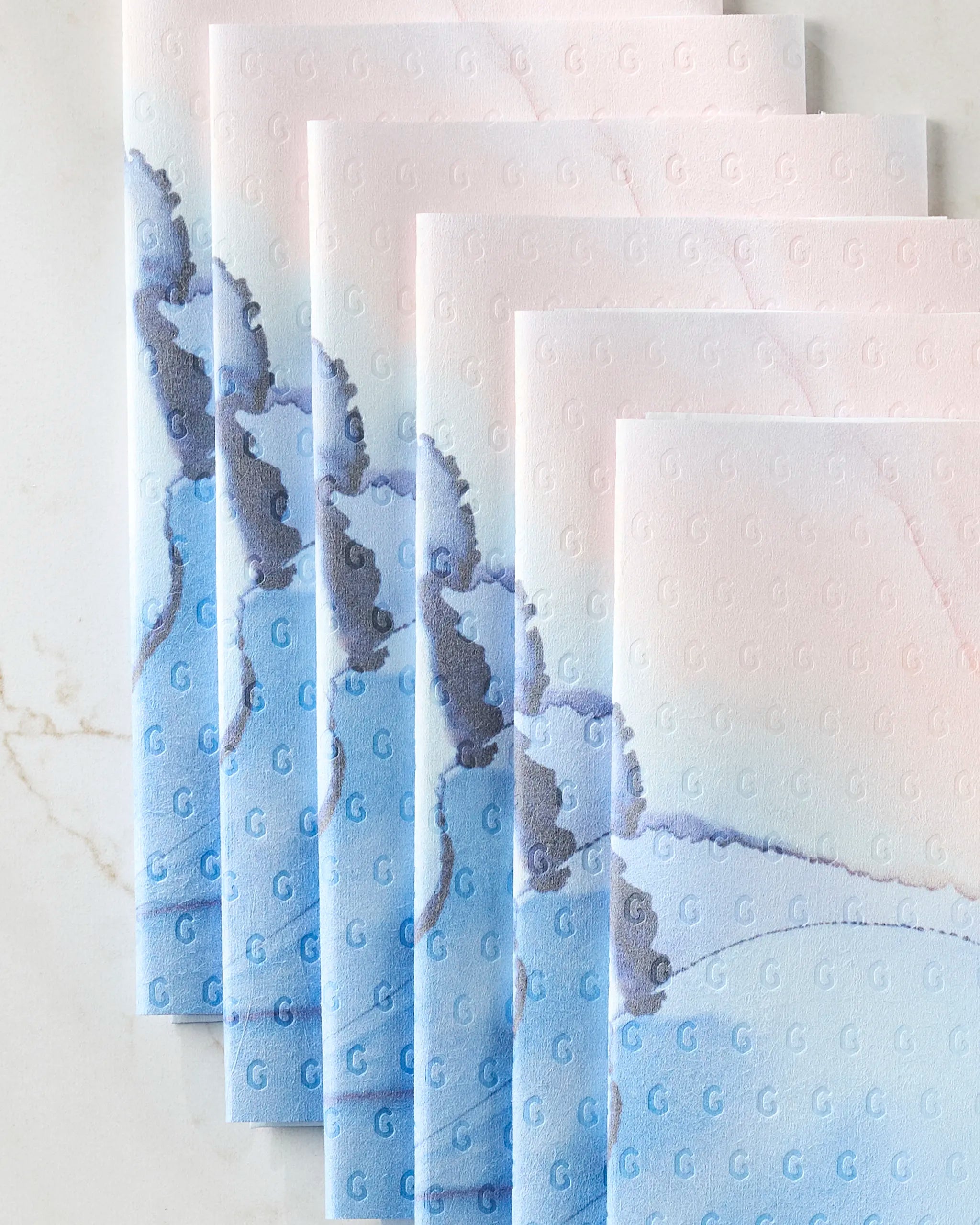 Five pastel-colored, eco-friendly Water And Ink paper towels by GEOMETRY featuring a blue and gray marbled design and an embossed “G” pattern are fanned out on a white marble surface.