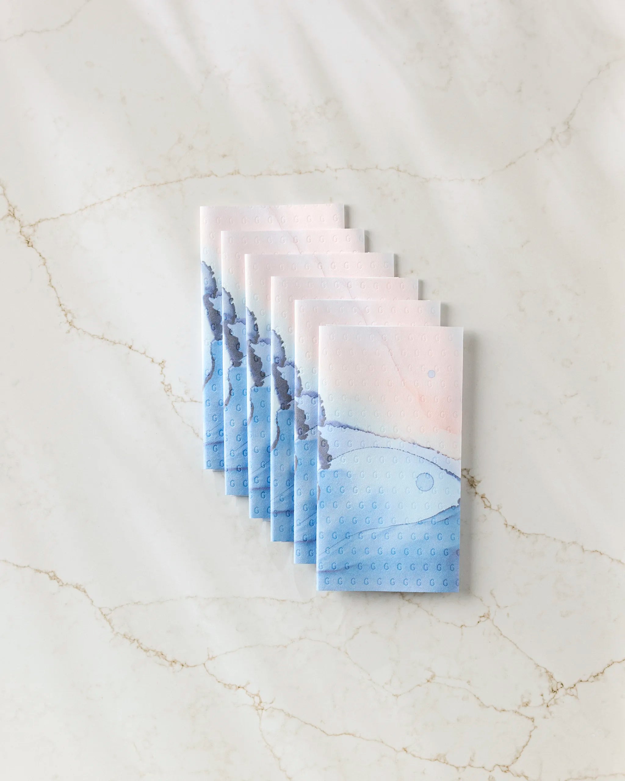Seven rectangular packets of GEOMETRY Water And Ink eco-friendly paper towels, featuring a blue and pink abstract design, are neatly fanned out on a white marble surface with light brown veining.