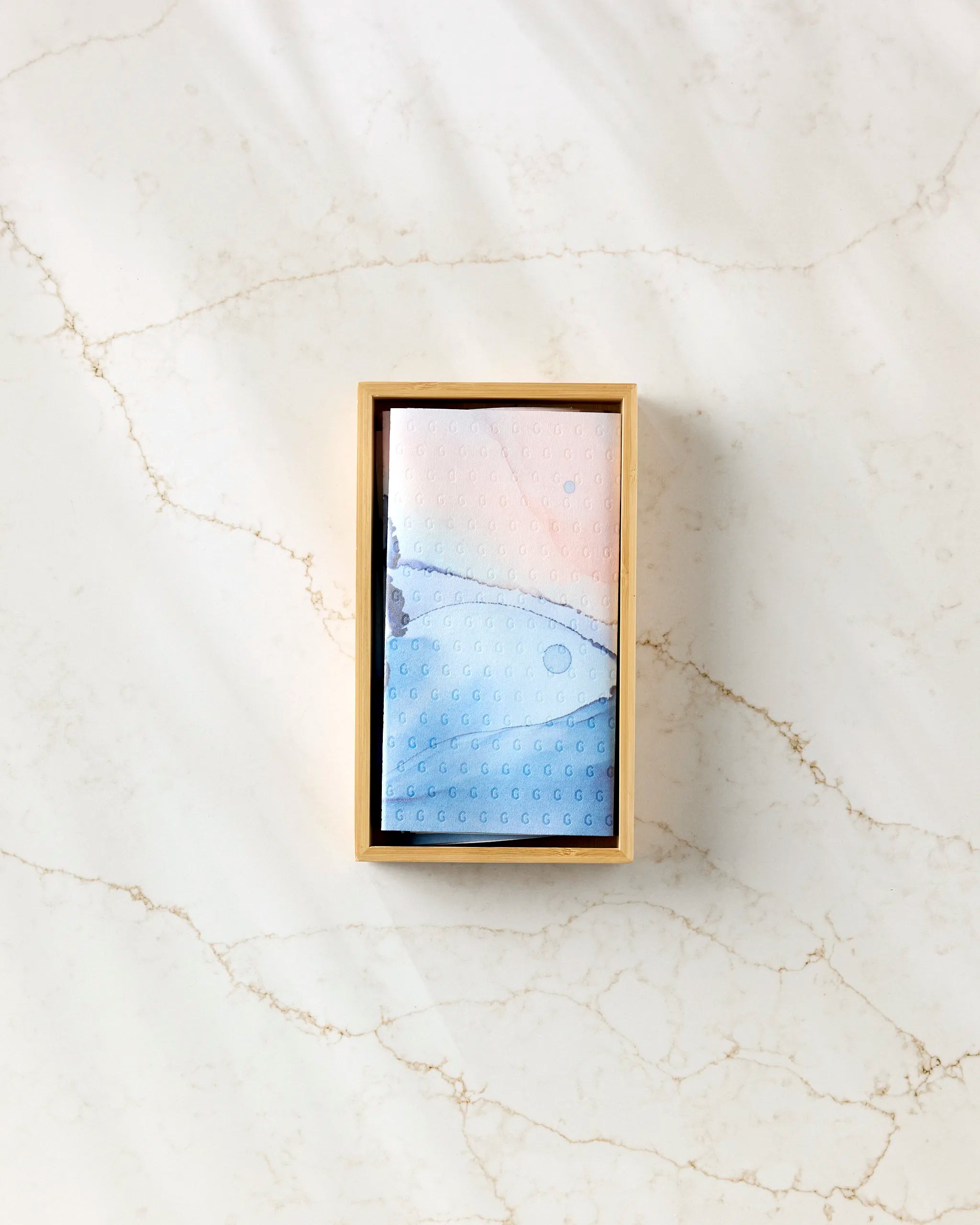 A rectangular wooden box holds "Water And Ink" by GEOMETRY, a blue and pink patterned item, on a subtly veined marble surface.