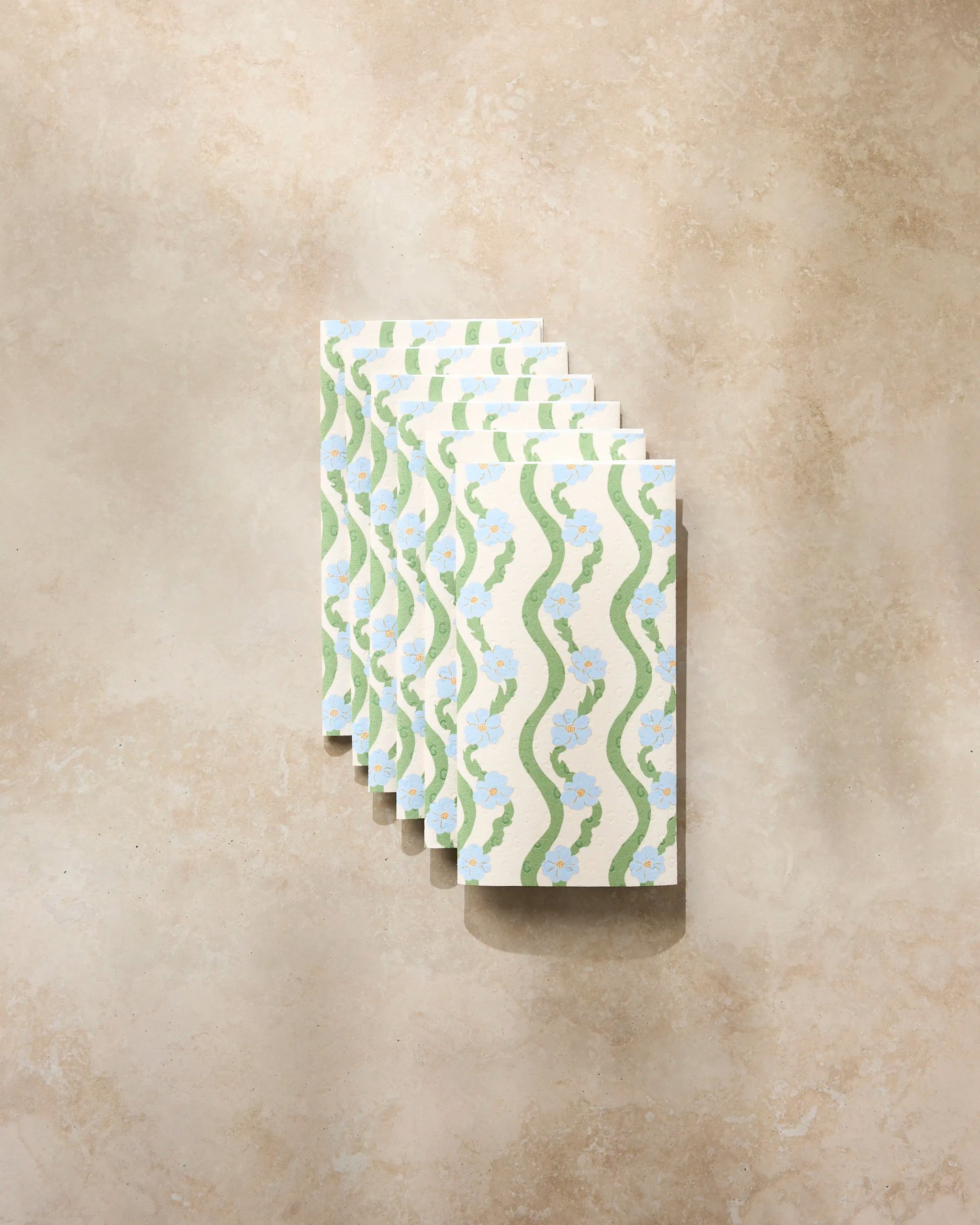 Six "Wavy Flowers" envelopes by Geometry, featuring green wavy lines and blue floral patterns, are arranged in a tidy stack on a textured light beige surface.