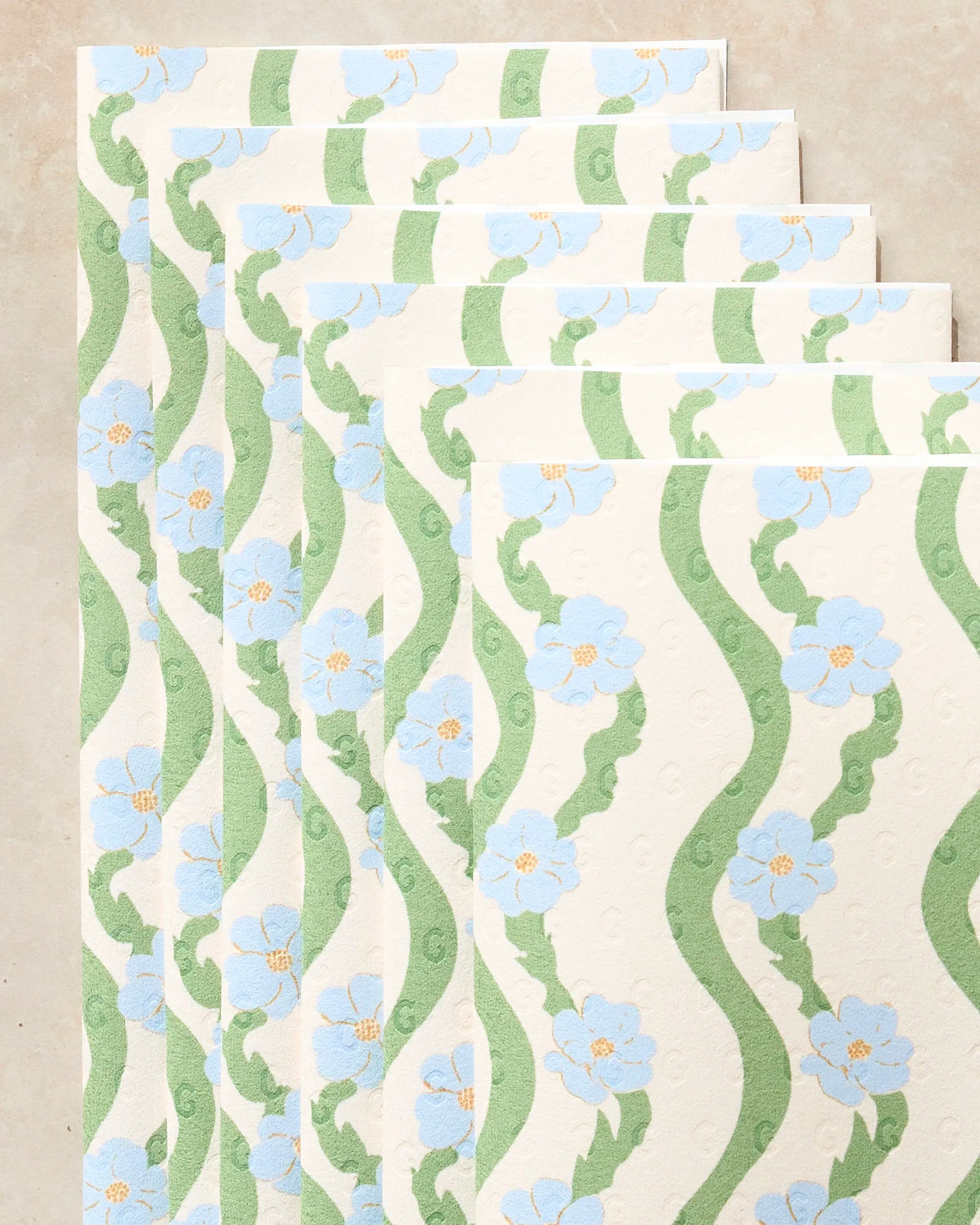 Five eco-friendly "Wavy Flowers" kitchen towels by Geometry, featuring green wavy lines and blue flowers, are arranged in a staggered fan shape on a light beige surface.