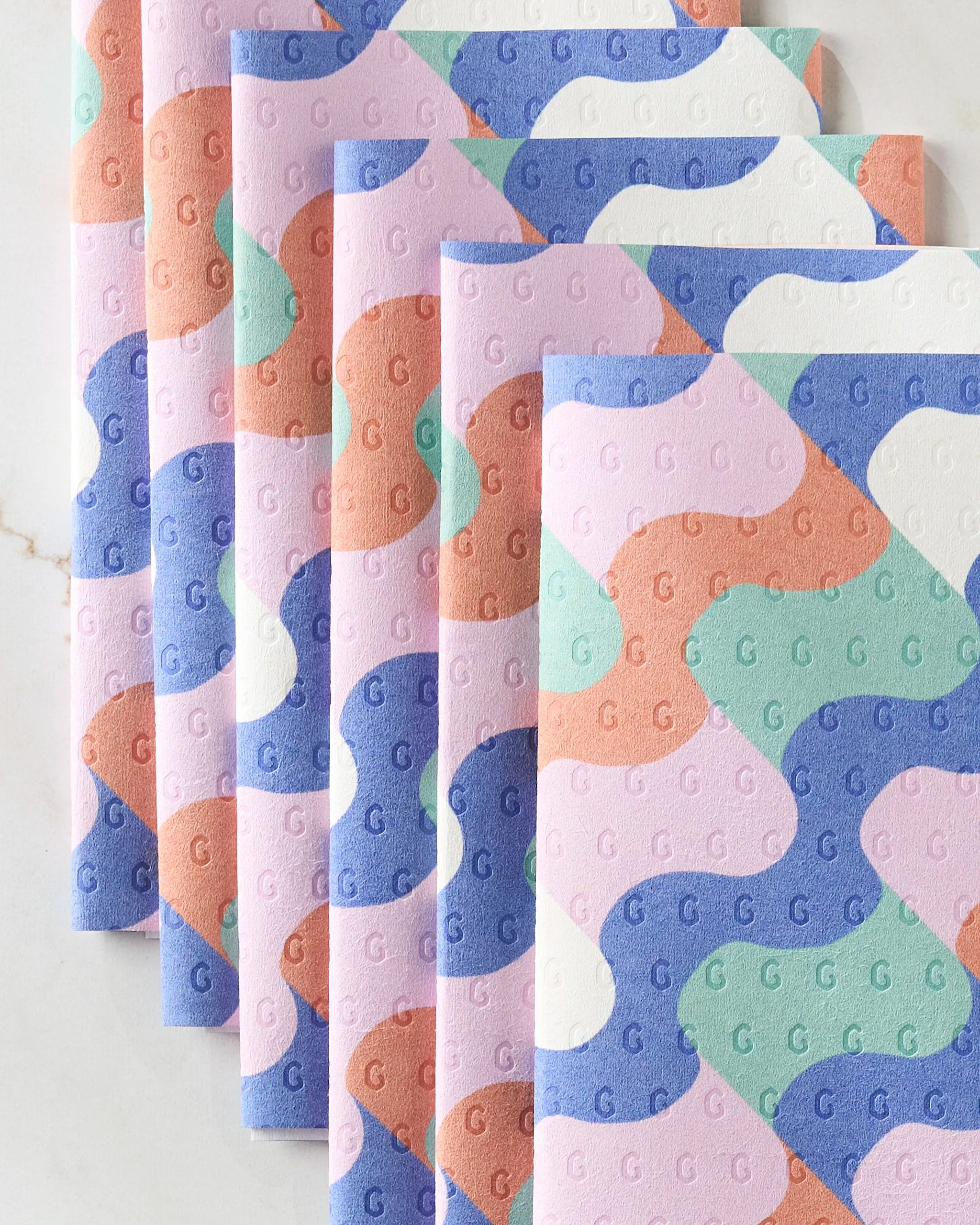 Claire Iglesias’s Wavy Stripes features five eco-friendly pastel paper towels in pink, blue, mint green, purple, and orange with abstract wavy patterns and embossed Gs, arranged fan-like on a light marble surface.