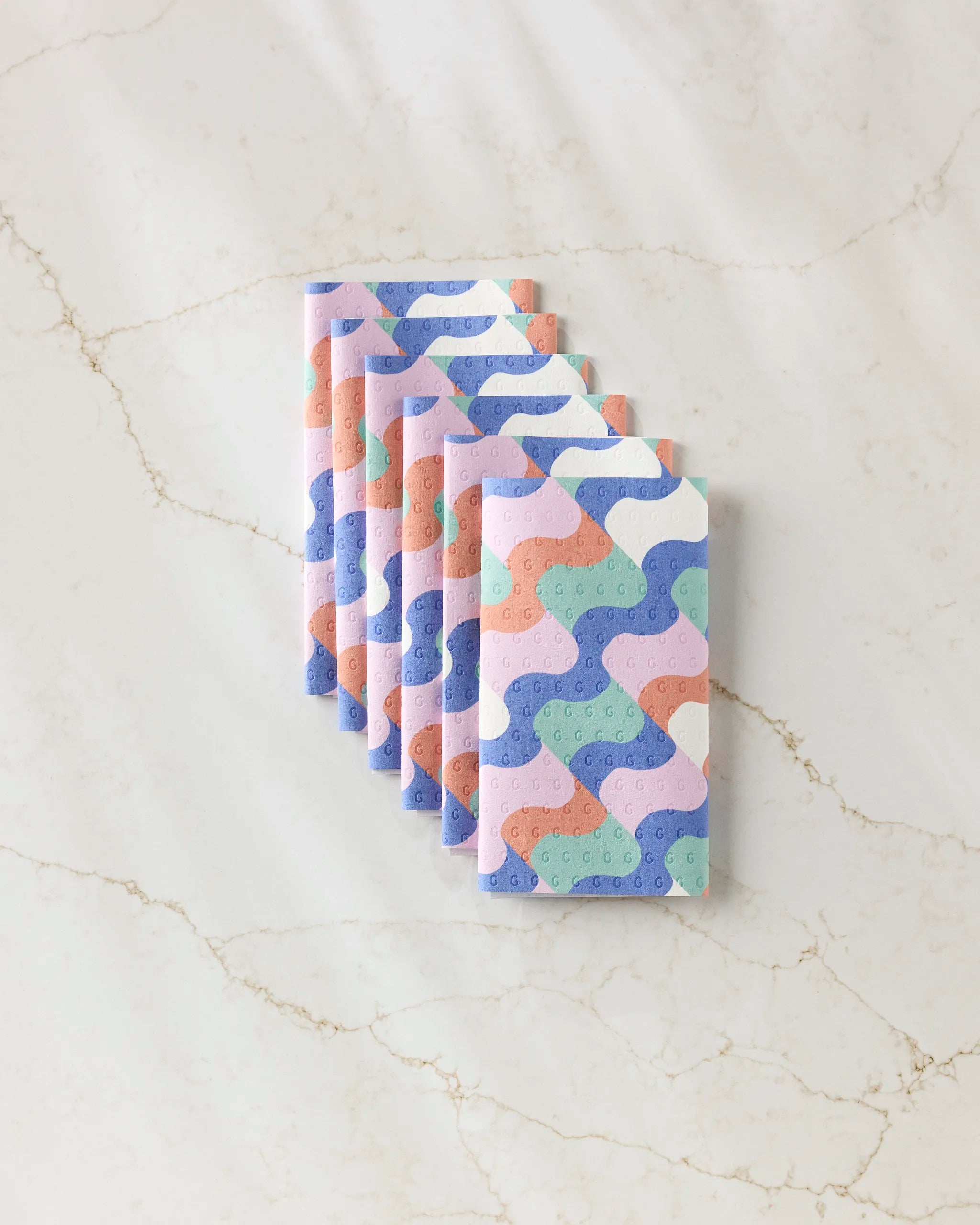 Six pastel notebooks from Claire Iglesias’s Wavy Stripes collection are fanned out on a white marble surface, featuring soft wavy designs in blue, pink, green, and purple for an eco-friendly look.