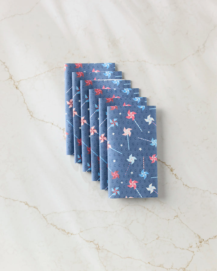 Six Whirlwind Pins blue napkins by Rebecca Bobko, featuring red and light blue pinwheels, are neatly arranged on a light marble surface—an eco-friendly alternative to disposable paper towels.