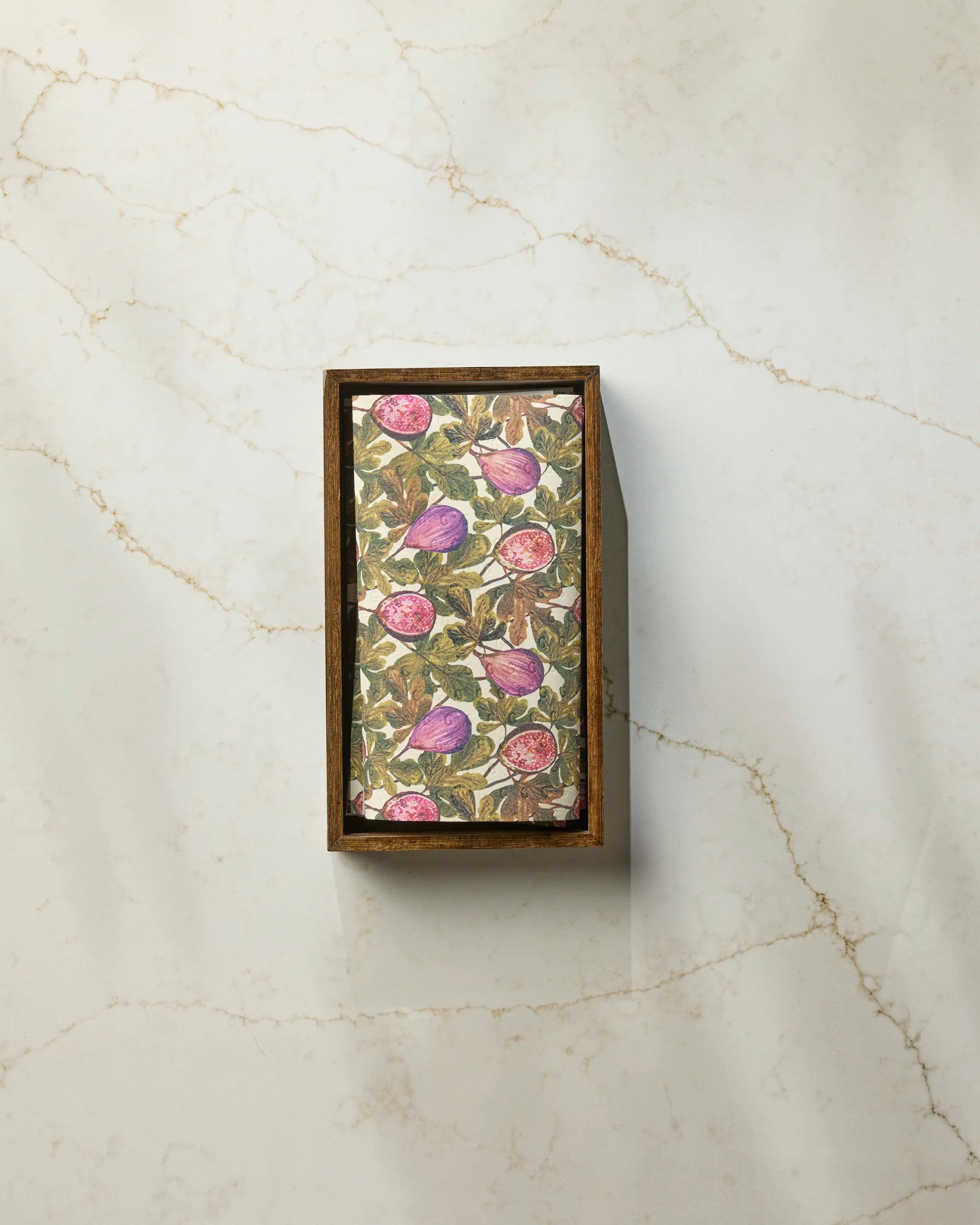 A rectangular Wild Fig bar soap by Geometry, wrapped in floral paper with purple and pink fruit designs, sits in a wooden box on light marble veined brown, paired with eco-friendly paper towels for a sustainable touch.