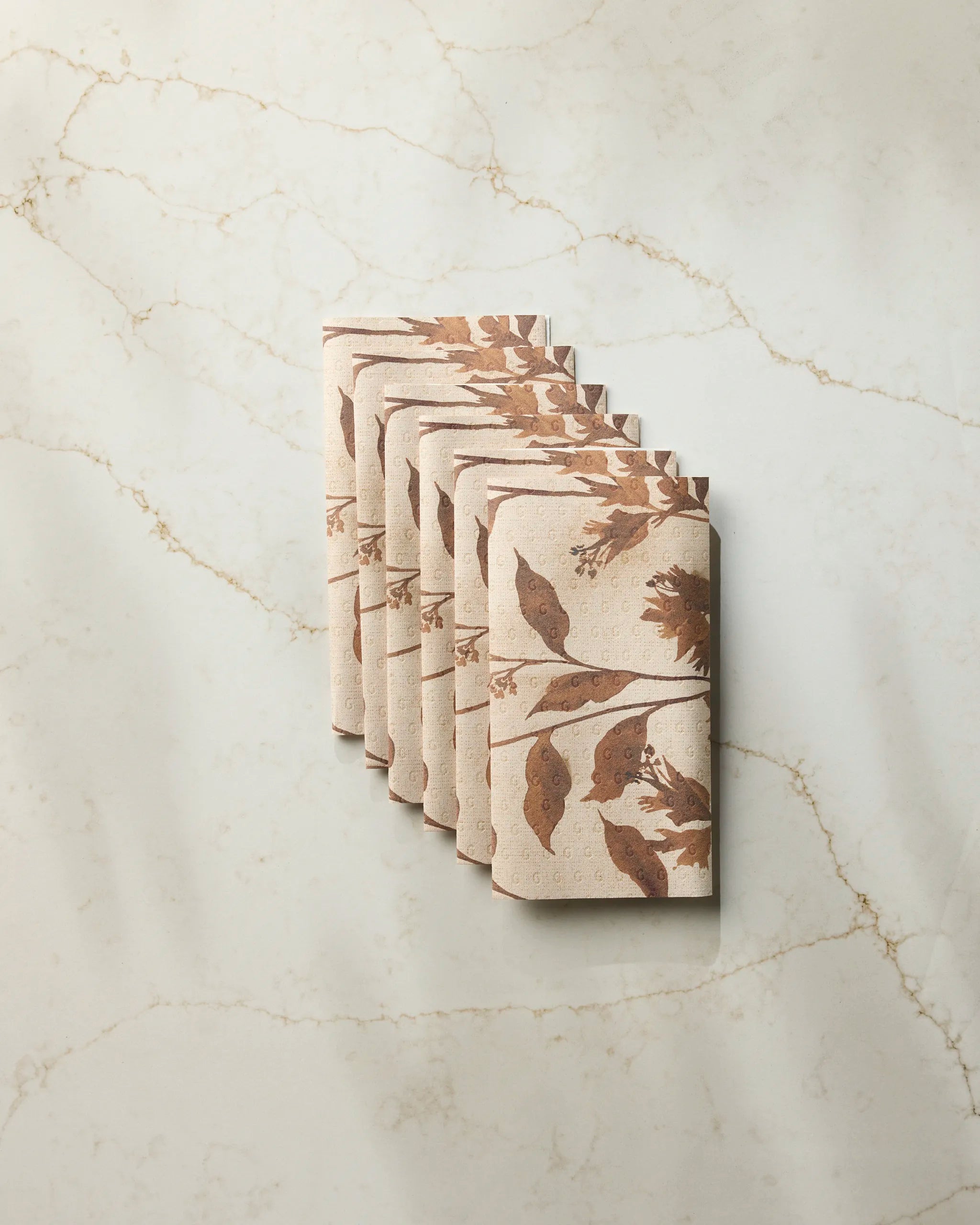 Six rolls of Willowmere toilet paper by Geometry, featuring a brown leaf and bird pattern on beige eco-friendly paper, are neatly stacked against a light marble wall with tan veins.