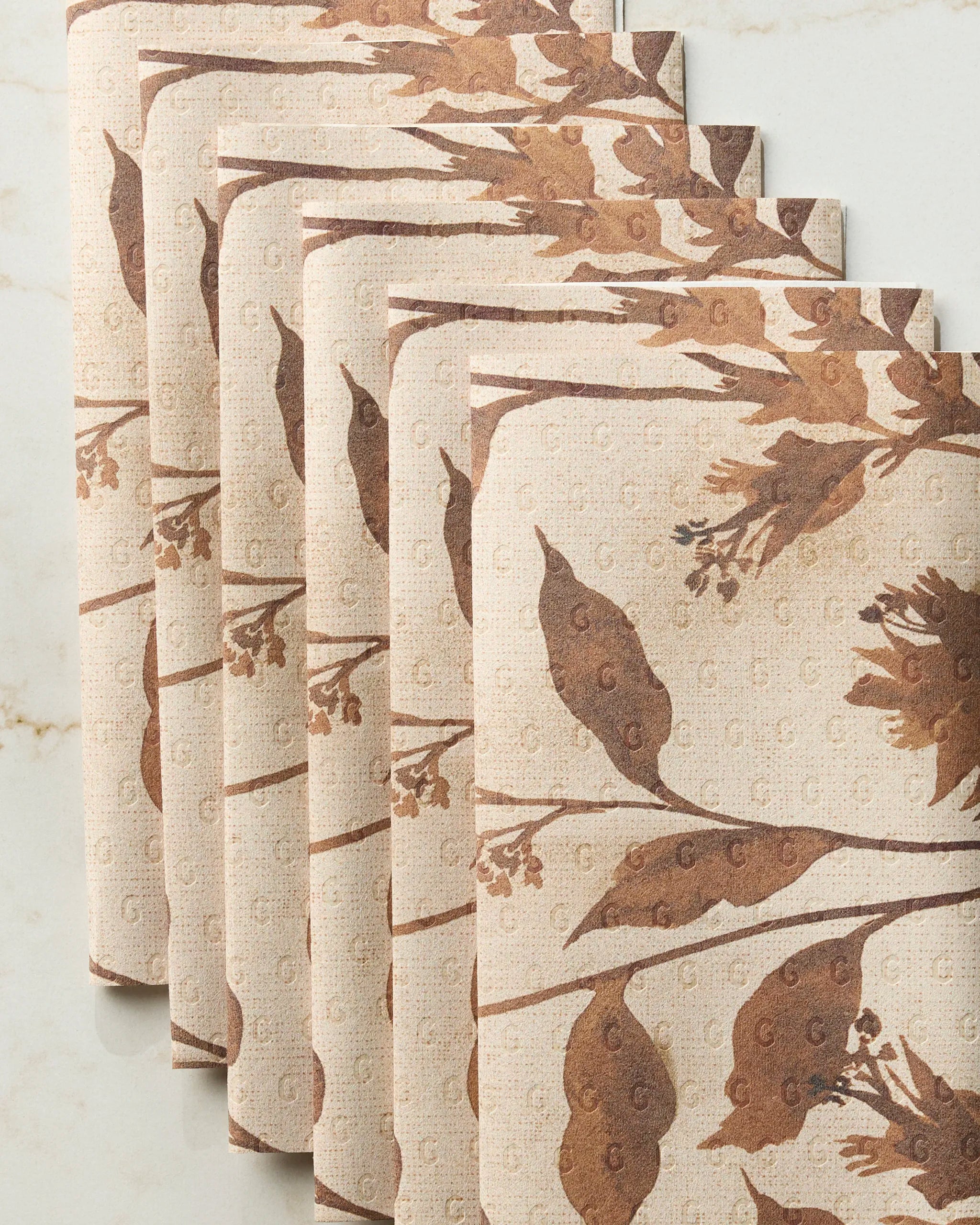 Five folded Willowmere napkins by Geometry, featuring a brown botanical leaf and branch pattern on beige, are neatly stacked on a light marble surface.