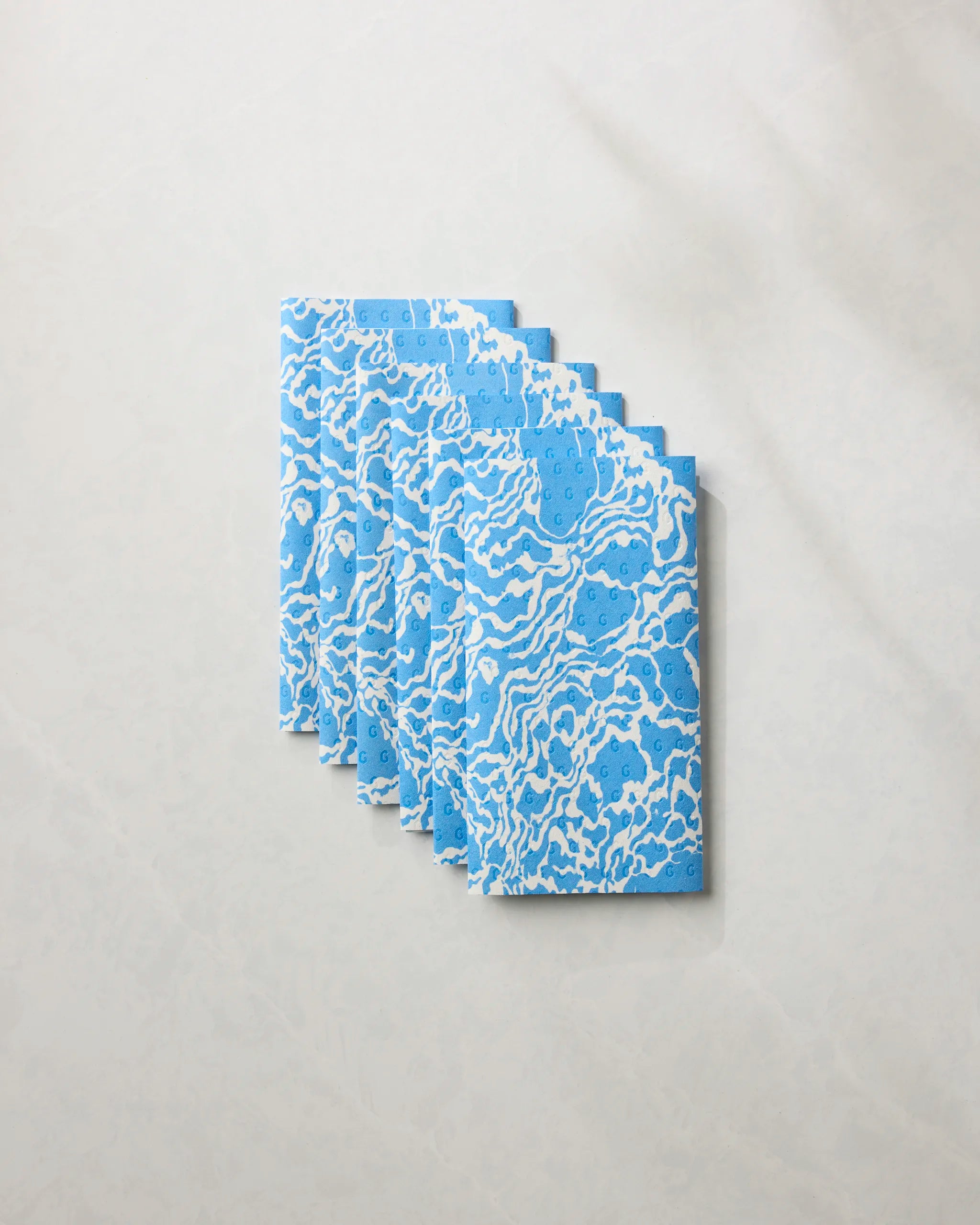Six Windward notebooks by Geometry, featuring a white abstract pattern on blue covers, are arranged in a fanned stack on a marble-like surface—perfect for eco-friendly paper towel users seeking stylish organization.
