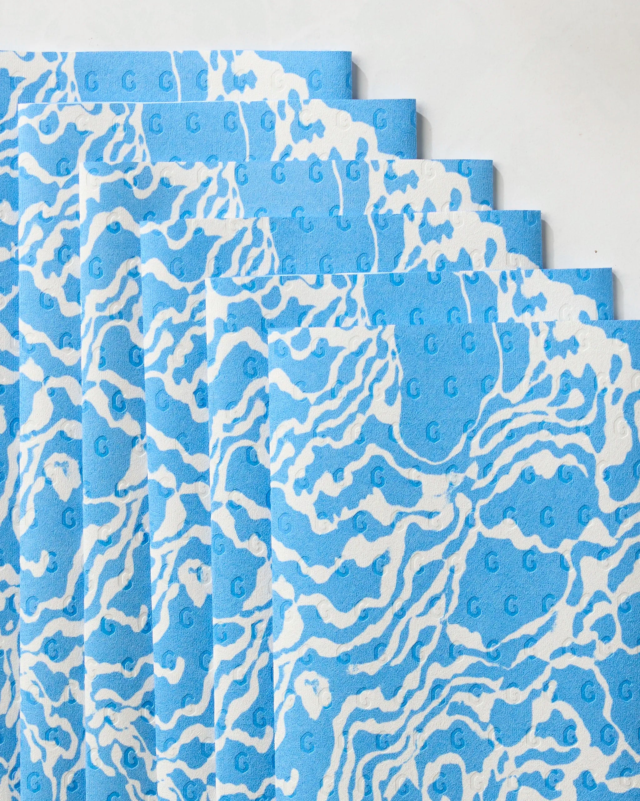 Six eco-friendly Geometry Windward paper towels, featuring a blue and white wavy abstract design with repeating G embossing, are fanned out on a white surface.