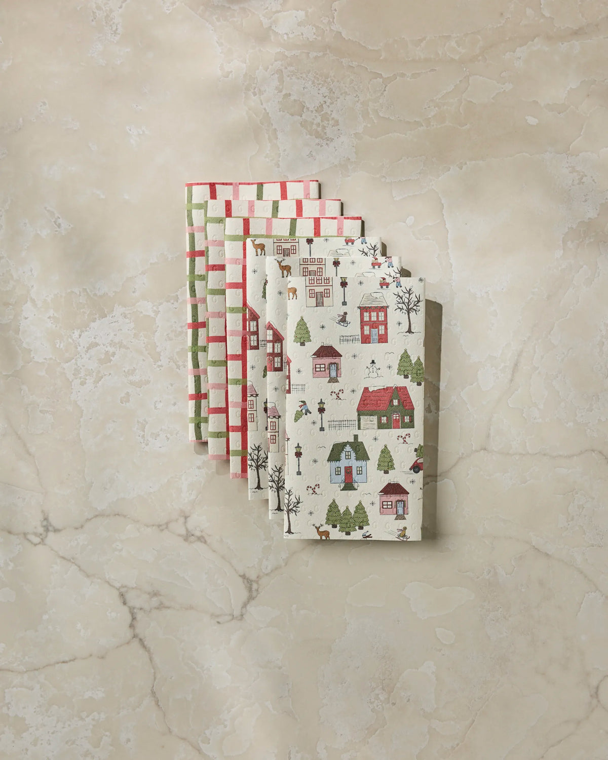 Four folded Geometry reusable paper towels in the Winter Village design are stacked on a marble surface. Two feature a red and white checkered pattern, while the other two display festive houses, trees, and holiday decorations in red, green, and cream tones.