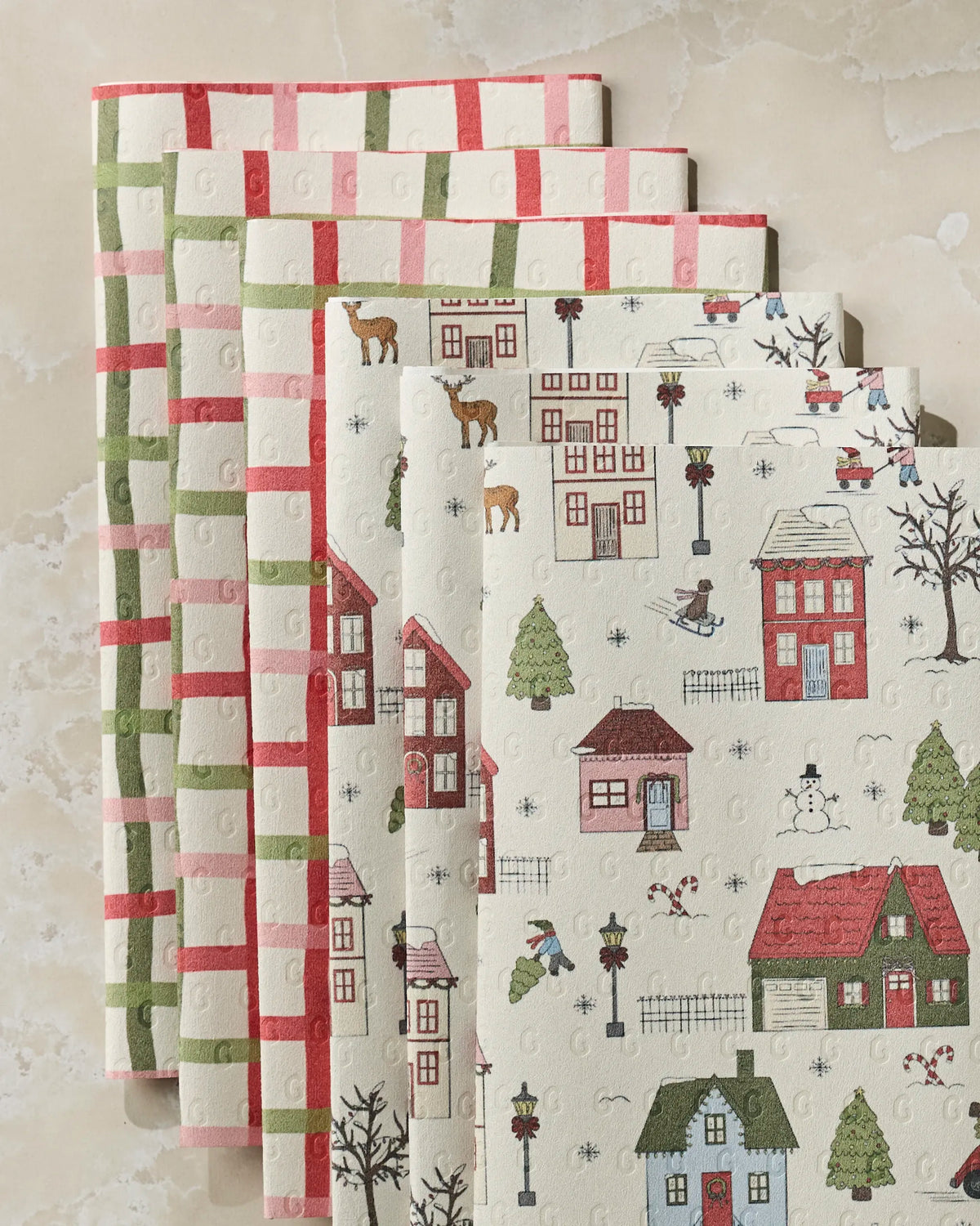 Five kitchen towels from Geometry—three featuring the Winter Village pattern and two with festive red and green checkered designs—are neatly stacked on a light-colored surface, making them an ideal choice for anyone looking to replace disposable options with reusable paper towels.