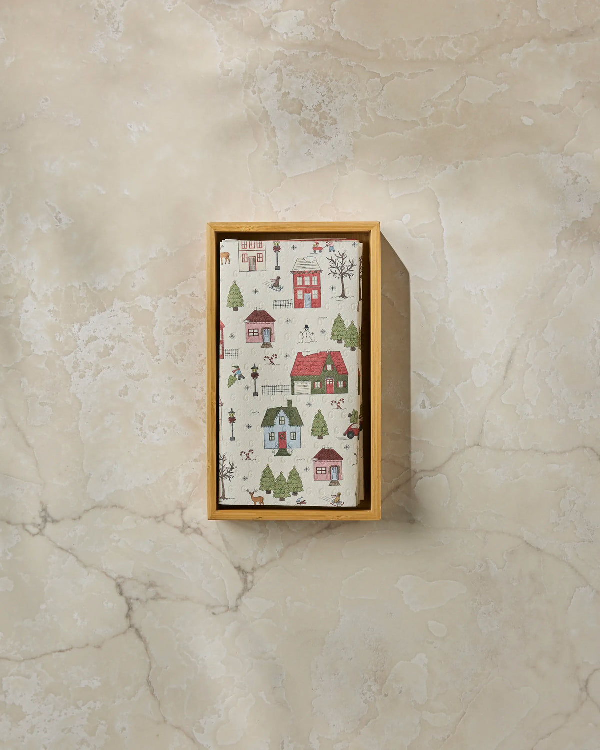 A rectangular box from Geometry called “Winter Village” sits on a light marble surface—its lid adorned with a charming village scene of small houses, trees, and people—making it perfect for storing your eco-friendly paper towels.